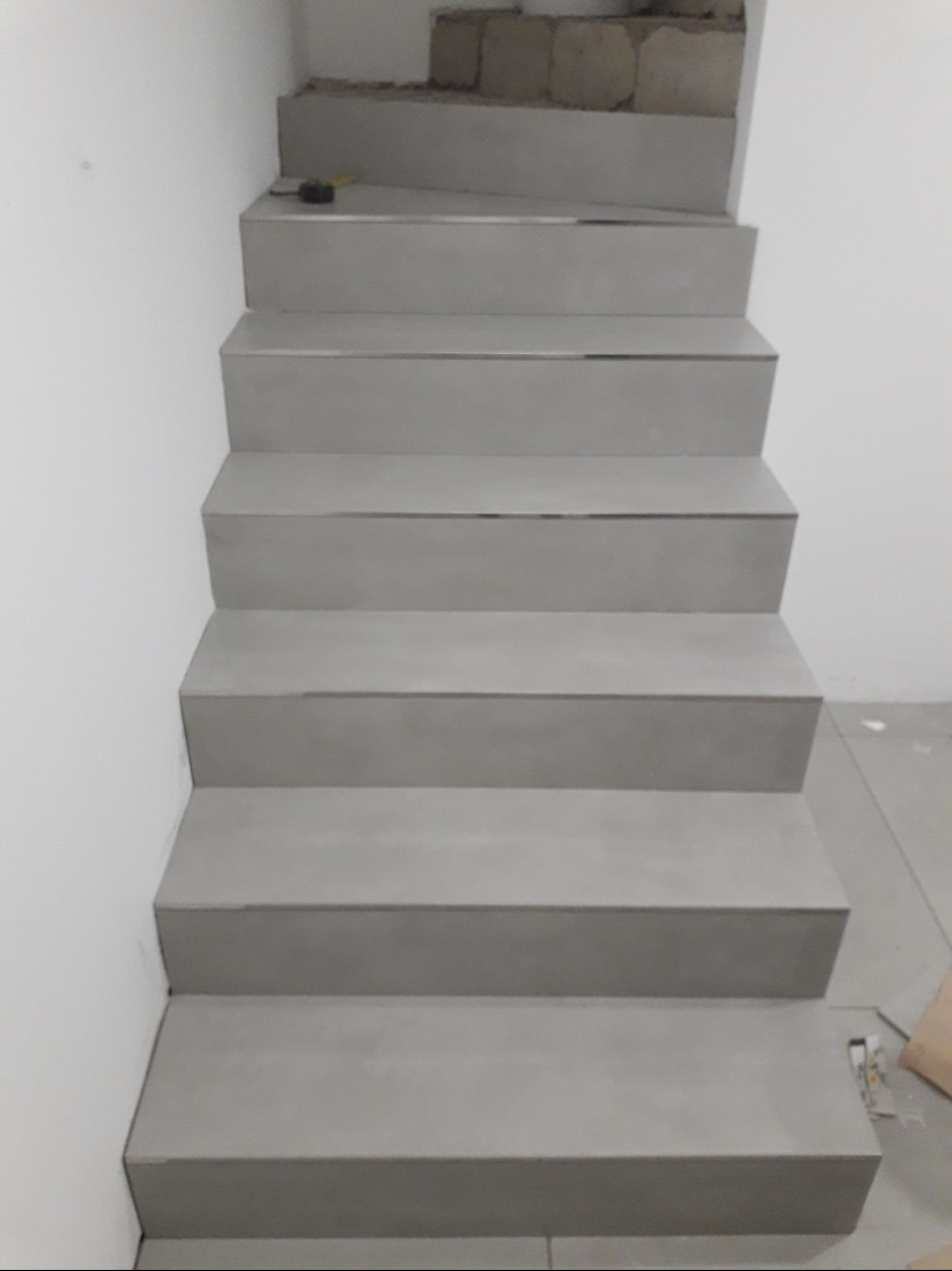 Schody wyłożone szarymi płytkami, widok z góry. Na pierwszym stopniu leży miarka. Część ściany i podłogi widoczna w tle. Minimalistyczny design.