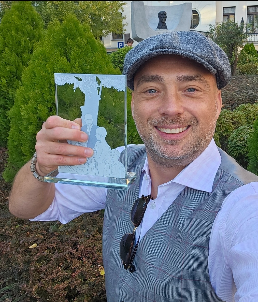 Mężczyzna w kaszkiecie prezentuje szklaną statuetkę z wygrawerowanym portretem rodziny na tle zieleni.