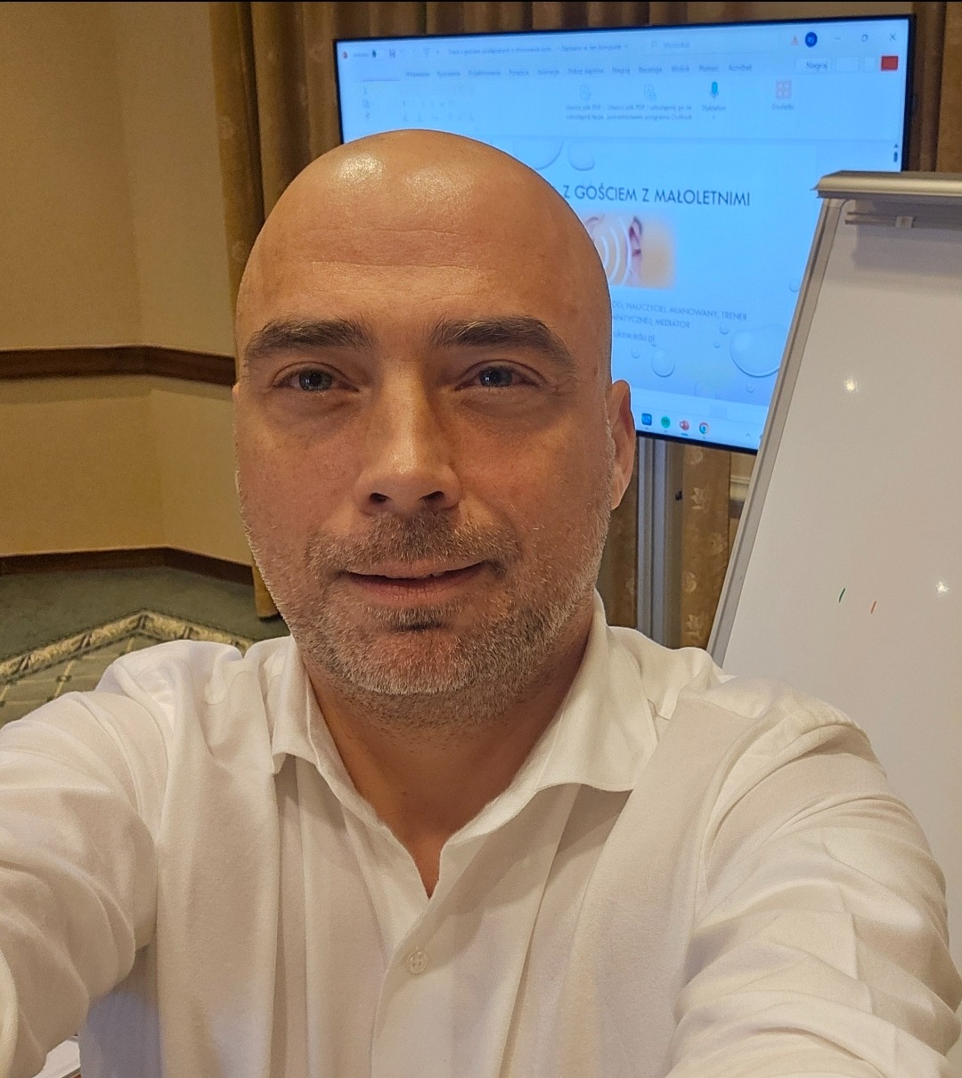 Portret mężczyzny w białej koszuli na tle ekranu z prezentacją o tematyce relacji z młodzieżą i tablicy flipchart. Widoczny uśmiech i kontakt wzrokowy.