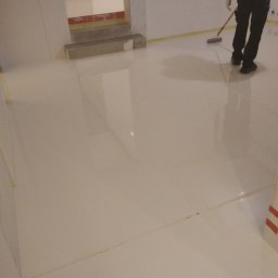 SIMEX - Aplikacja posadzki żywicznej: pracownik w czarnych spodniach i niebieskiej koszulce rozprowadza żywicę za pomocą mopa na długim kiju, widoczna gładka, błyszcząca powierzchnia i żółta linia wzdłuż...