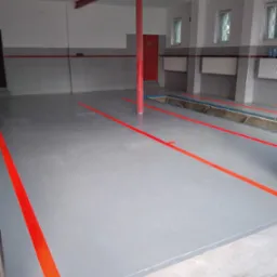 Szara posadzka w garażu z wyznaczonymi pomarańczowymi liniami, widoczne kanały rewizyjne i czerwona kolumna podtrzymująca strop.