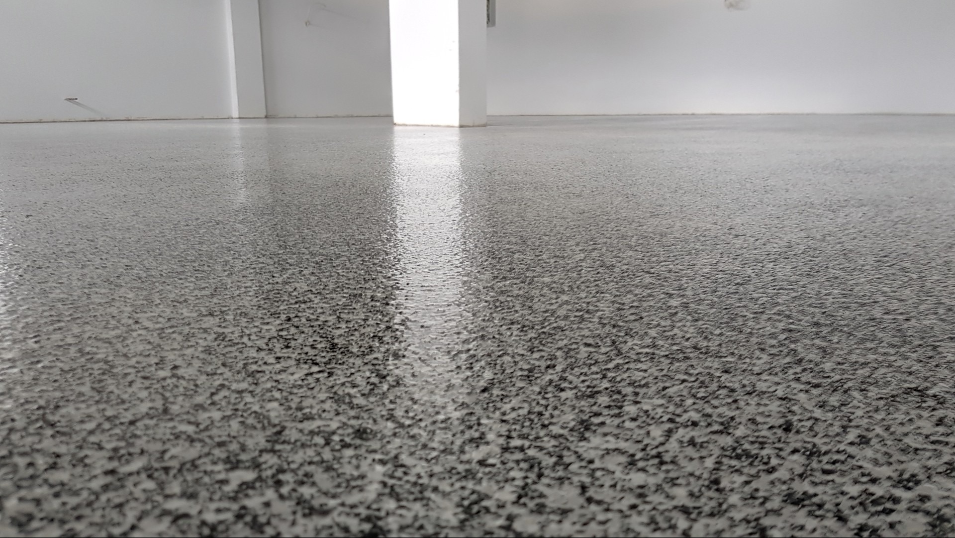 Świeżo wylana, błyszcząca podłoga z szarej żywicy epoksydowej z delikatnym wzorem drobnych, ciemnych kropek, odbijająca światło w pustym, białym pomieszczeniu z filarem.