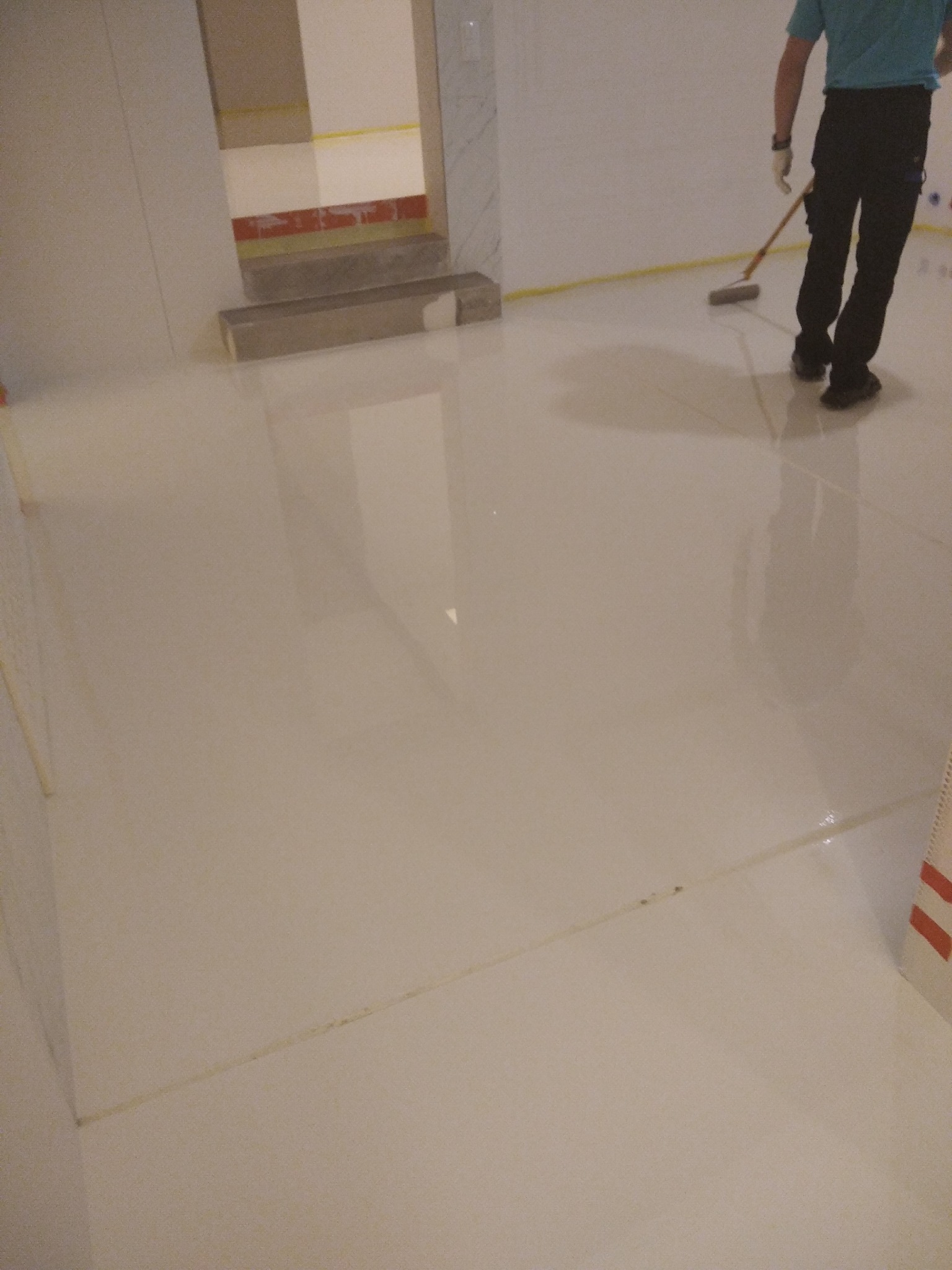 Aplikacja posadzki żywicznej: pracownik w czarnych spodniach i niebieskiej koszulce rozprowadza żywicę za pomocą mopa na długim kiju, widoczna gładka, błyszcząca powierzchnia i żółta linia wzdłuż...