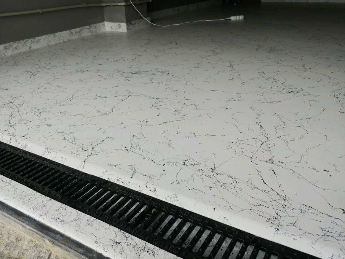 Jasna posadzka z ciemnymi żyłkowatymi wzorami, zakończona czarnym odpływem liniowym i listwą przypodłogową w tle.