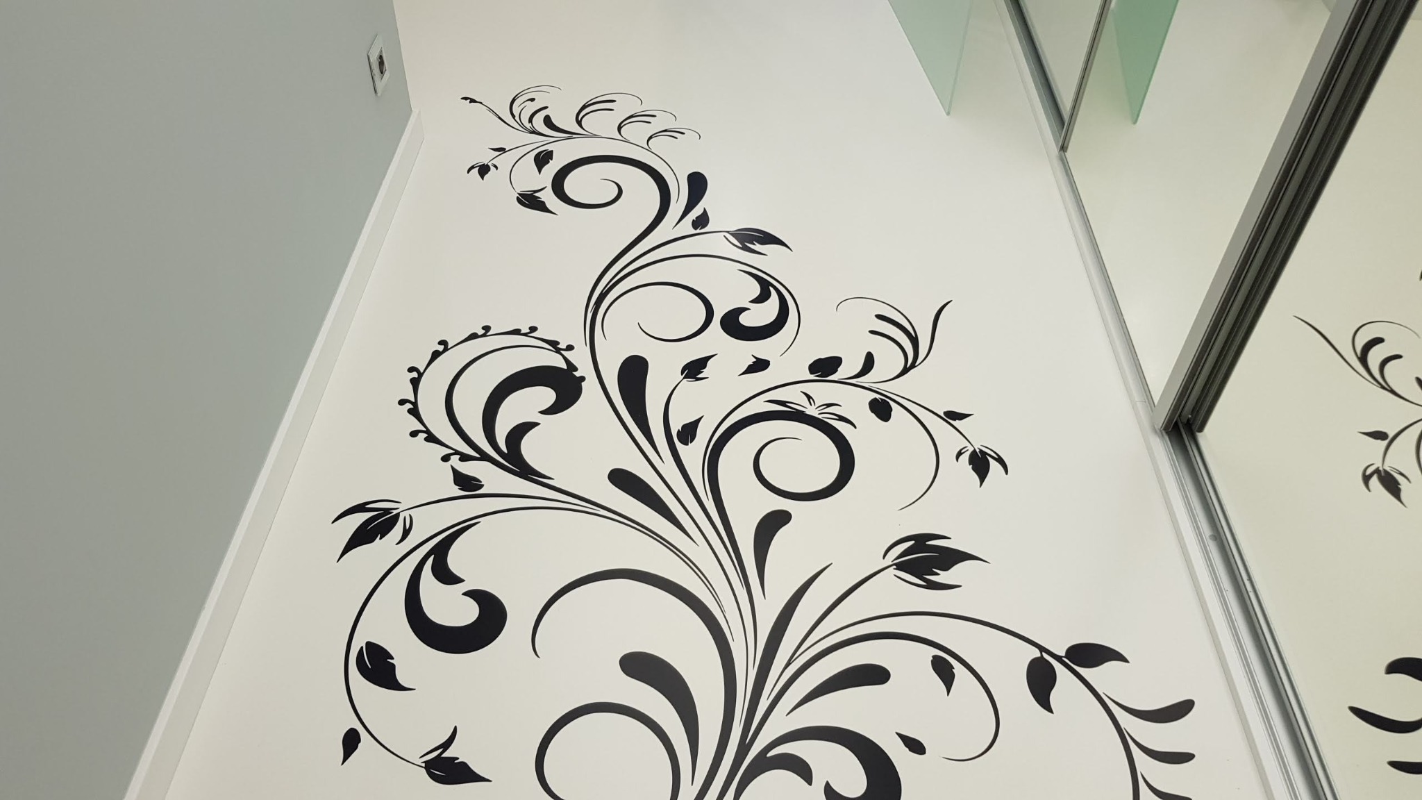 Czarny, roślinny ornament na białej ścianie w rogu pomieszczenia, obok lustrzanych drzwi szafy przesuwnej.