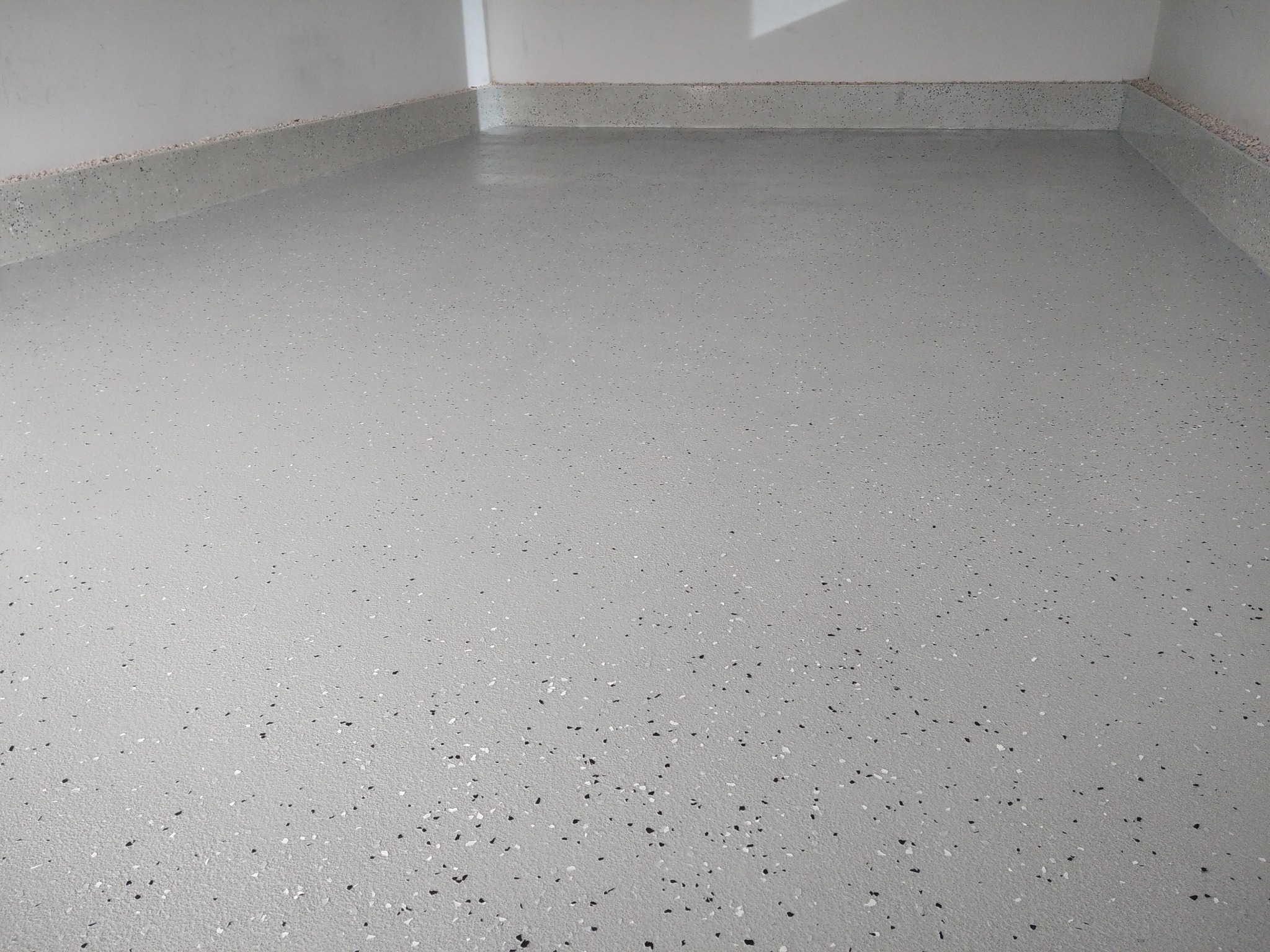 Jasnoszara, gładka posadzka z drobnymi, czarnymi i białymi plamkami, z delikatnym połyskiem, świeżo wylana, z widoczną listwą przypodłogową w tym samym kolorze.