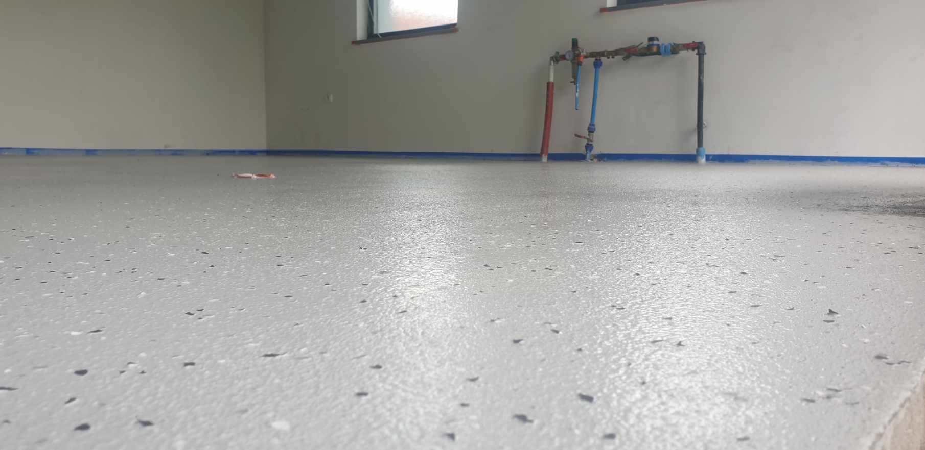 Świeżo wylana, jasnoszara posadzka z delikatnymi, ciemnymi plamkami, zabezpieczona niebieską taśmą malarską przy ścianach. W tle widoczne rury instalacyjne przy ścianie i okno.