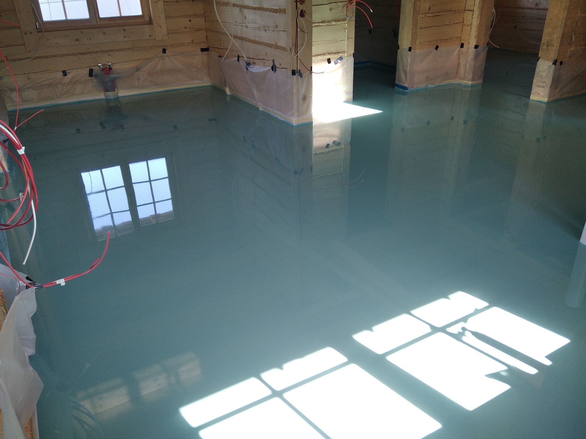 Świeżo wylana, samopoziomująca się posadzka w surowym wnętrzu drewnianego domu, zabezpieczonym folią malarską, z odbiciem okna w mokrej powierzchni.