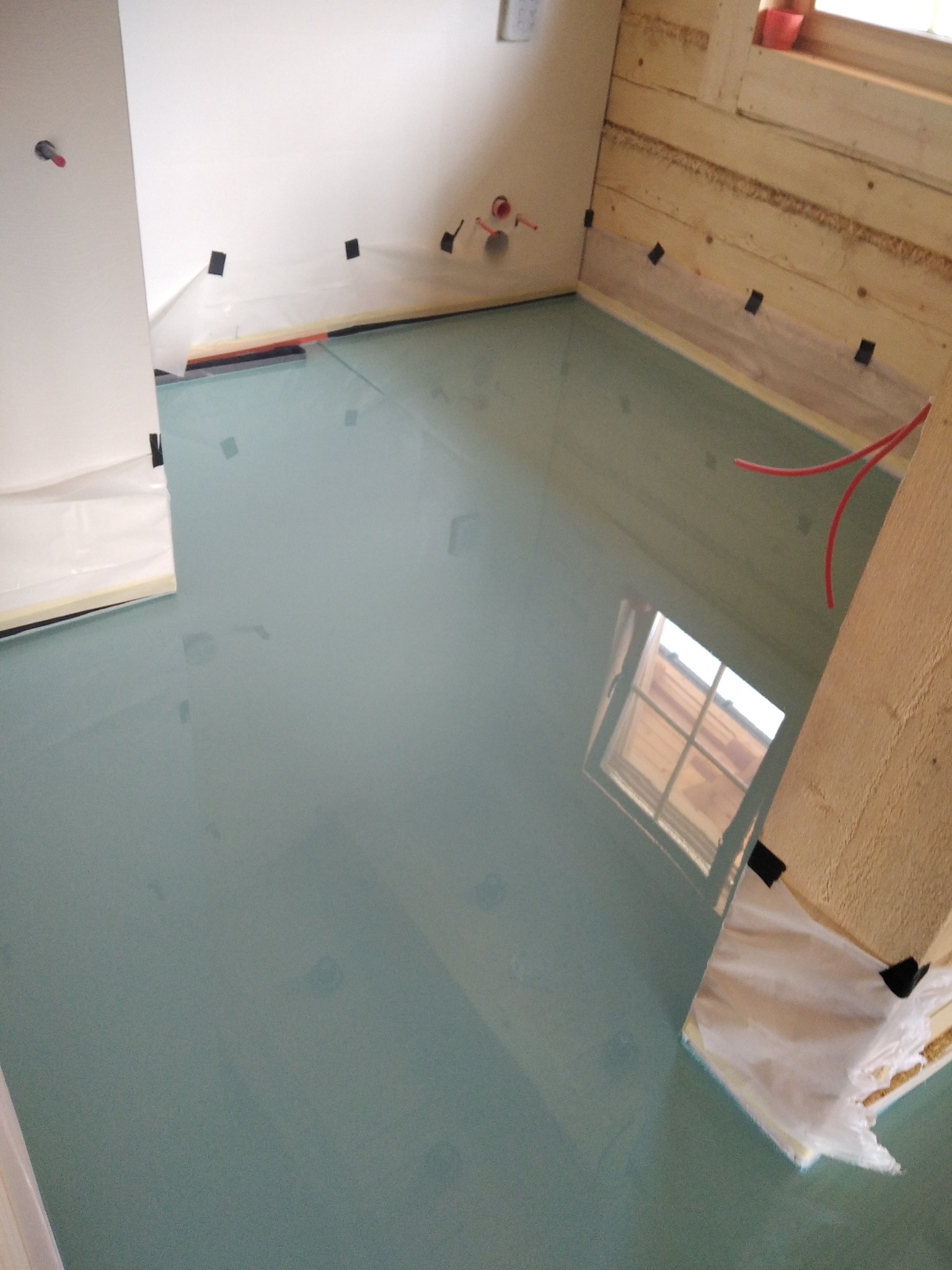 Świeżo wylana, jasnoniebieska posadzka w pomieszczeniu z surowymi drewnianymi ścianami, zabezpieczonymi folią i taśmą malarską, widoczne rury ogrzewania podłogowego.