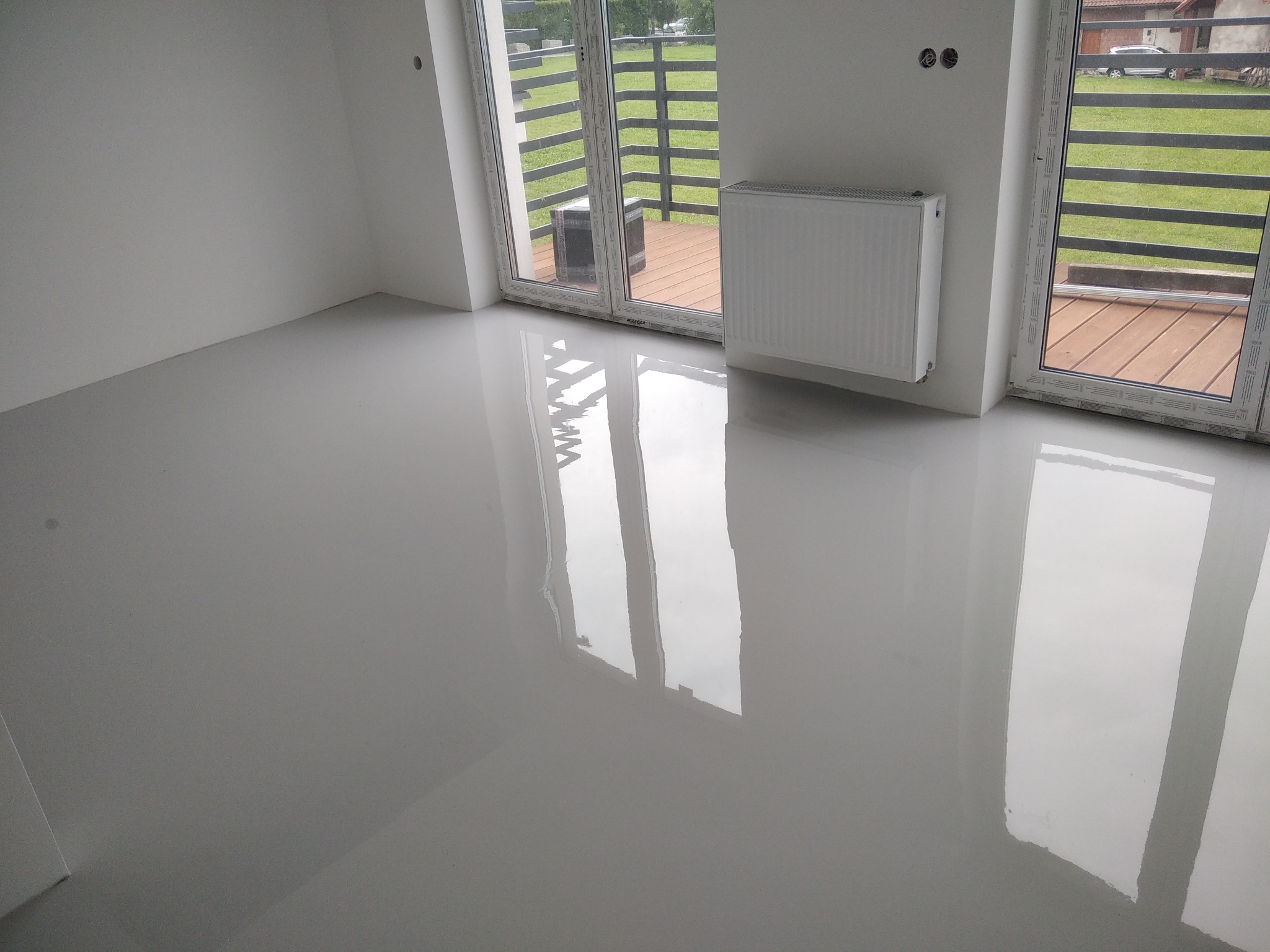 Jasnoszara posadzka żywiczna z połyskiem, odbijająca światło z okien balkonowych, z widocznym grzejnikiem na ścianie.