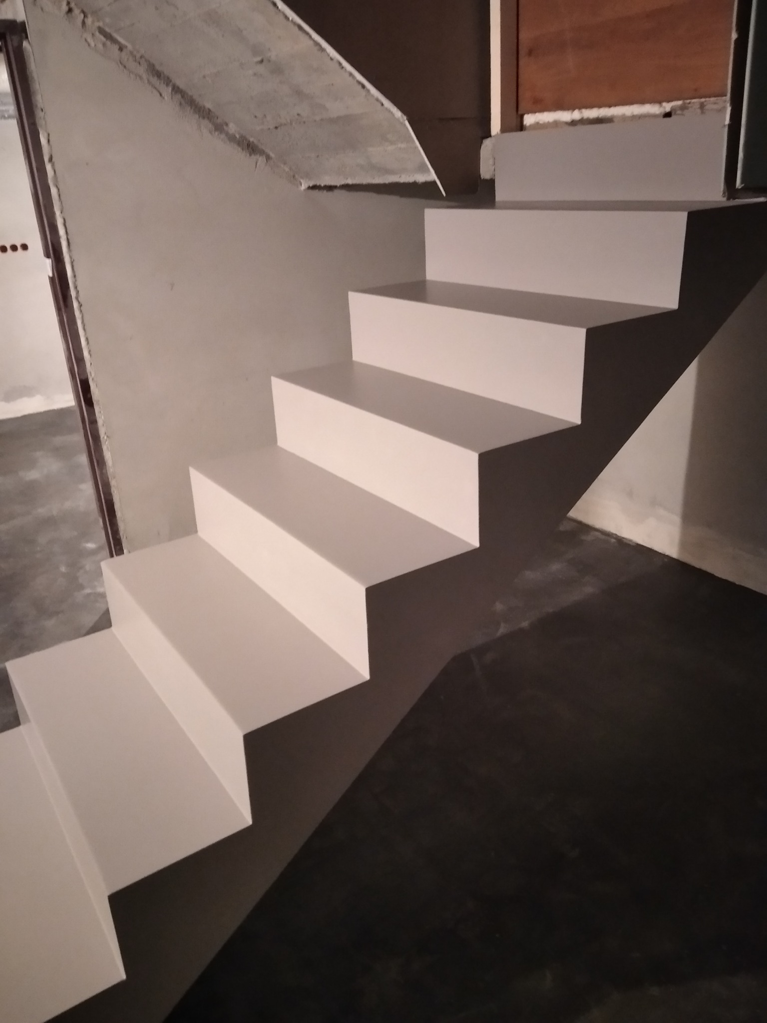 Białe, minimalistyczne schody w surowym wnętrzu, z widocznym betonowym stropem i ciemną podłogą, w trakcie prac wykończeniowych.