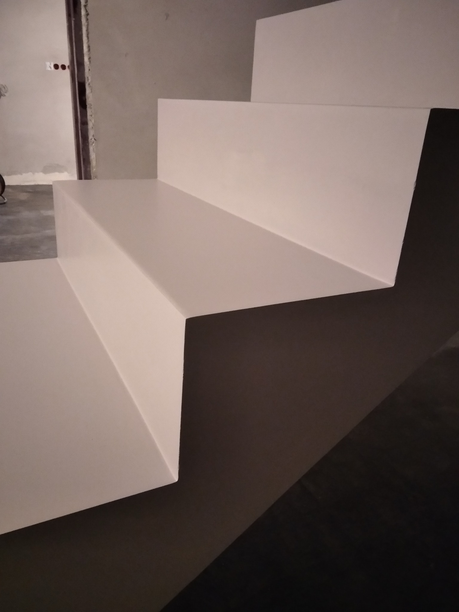 Nowoczesne, białe schody z gładką powierzchnią w surowym wnętrzu, z widocznym fragmentem ściany w stanie deweloperskim i ciemną podłogą.