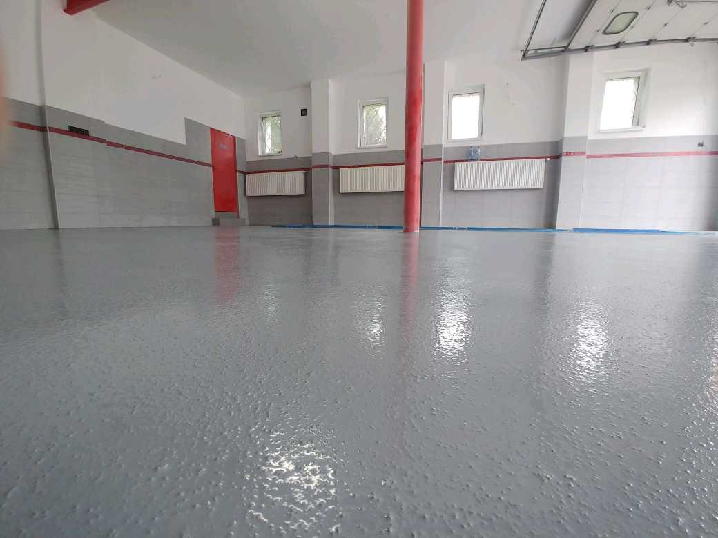 Szara, błyszcząca posadzka w przestronnym pomieszczeniu z czerwonym słupem i drzwiami, widoczne grzejniki i okna, ściany częściowo wyłożone szarymi płytkami.
