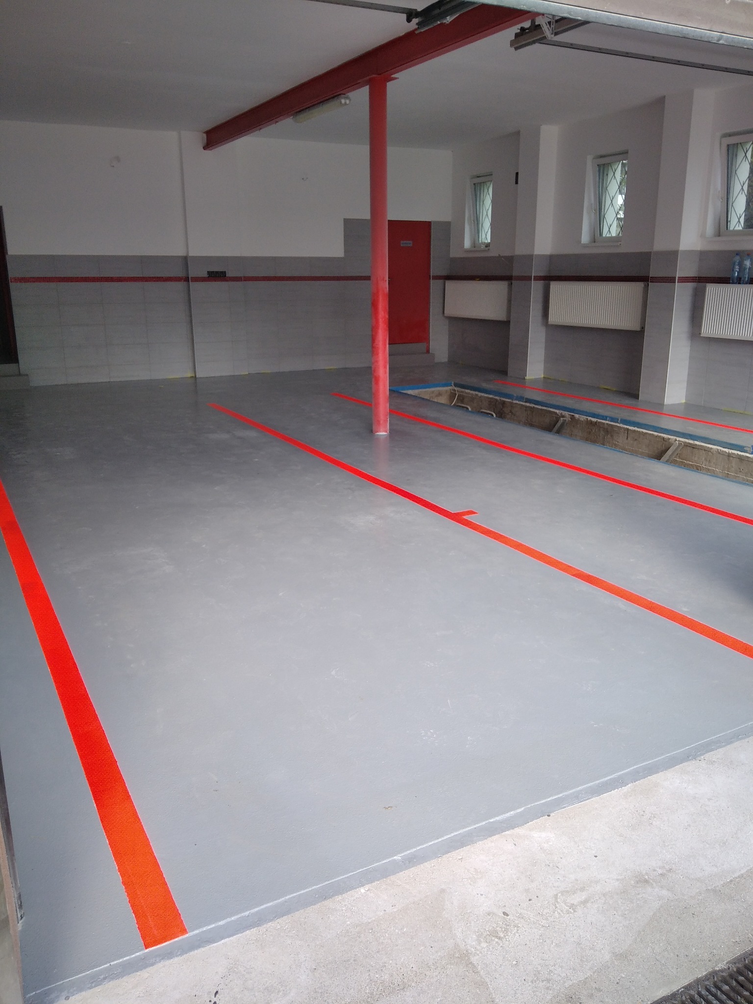 Szara posadzka w garażu z wyznaczonymi pomarańczowymi liniami, widoczne kanały rewizyjne i czerwona kolumna podtrzymująca strop.