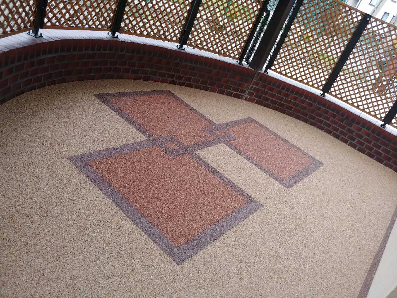 Balkon z beżową, kamyczkową posadzką i geometrycznym wzorem z brązowych prostokątów obramowanych ciemniejszym odcieniem, otoczony ceglaną balustradą i drewnianą kratką.