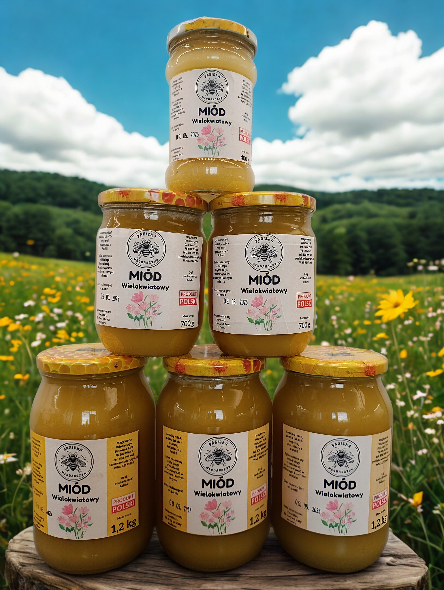 Piramida z słoików miodu wielokwiatowego na drewnianym pniu, na tle łąki i błękitnego nieba. Widoczne etykiety z nazwą pasieki i datą ważności.