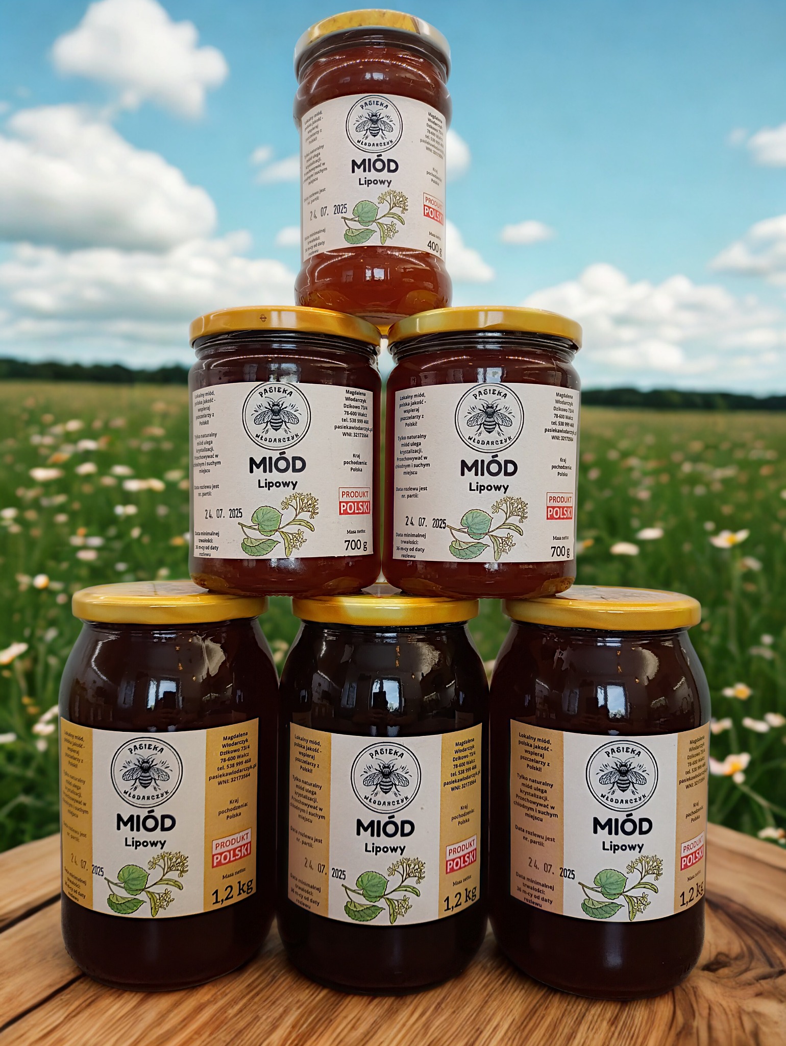 Piramida z sześciu słoików z miodem lipowym na drewnianym blacie, na tle łąki i błękitnego nieba. Widoczne etykiety z nazwą pasieki i informacjami o produkcie.