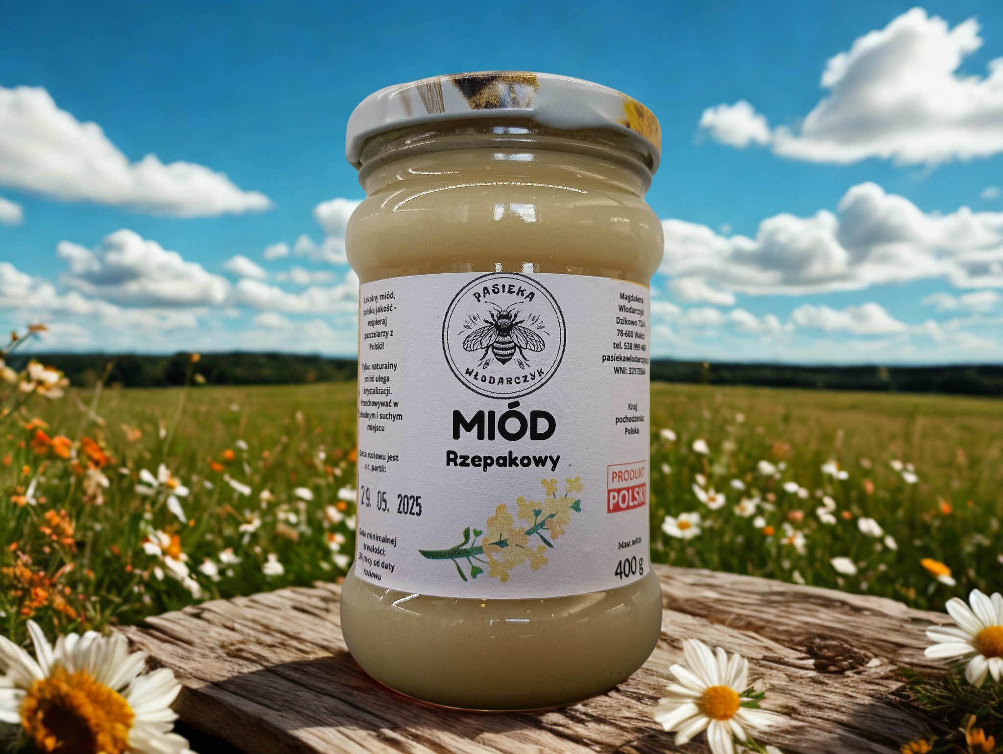 Słoik rzepakowego miodu z Pasieki Włodarczyk na tle łąki i błękitnego nieba. Słoik stoi na drewnianej desce, a wokół niego leżą białe stokrotki.