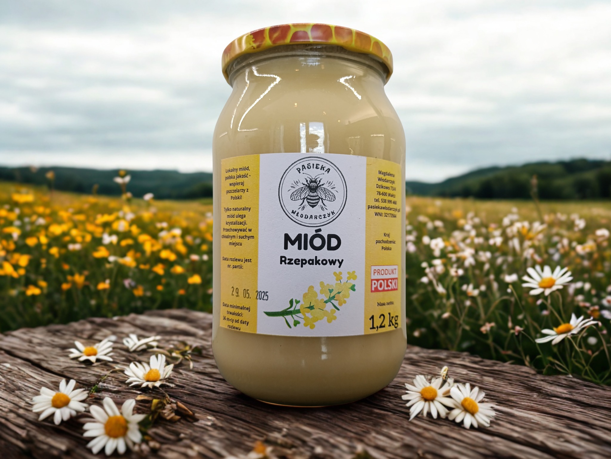 Słoik rzepakowego miodu z etykietą 'Pasieka Włodarczyk' na drewnianym stole, otoczony stokrotkami, z polem kwiatów i wzgórzami w tle.