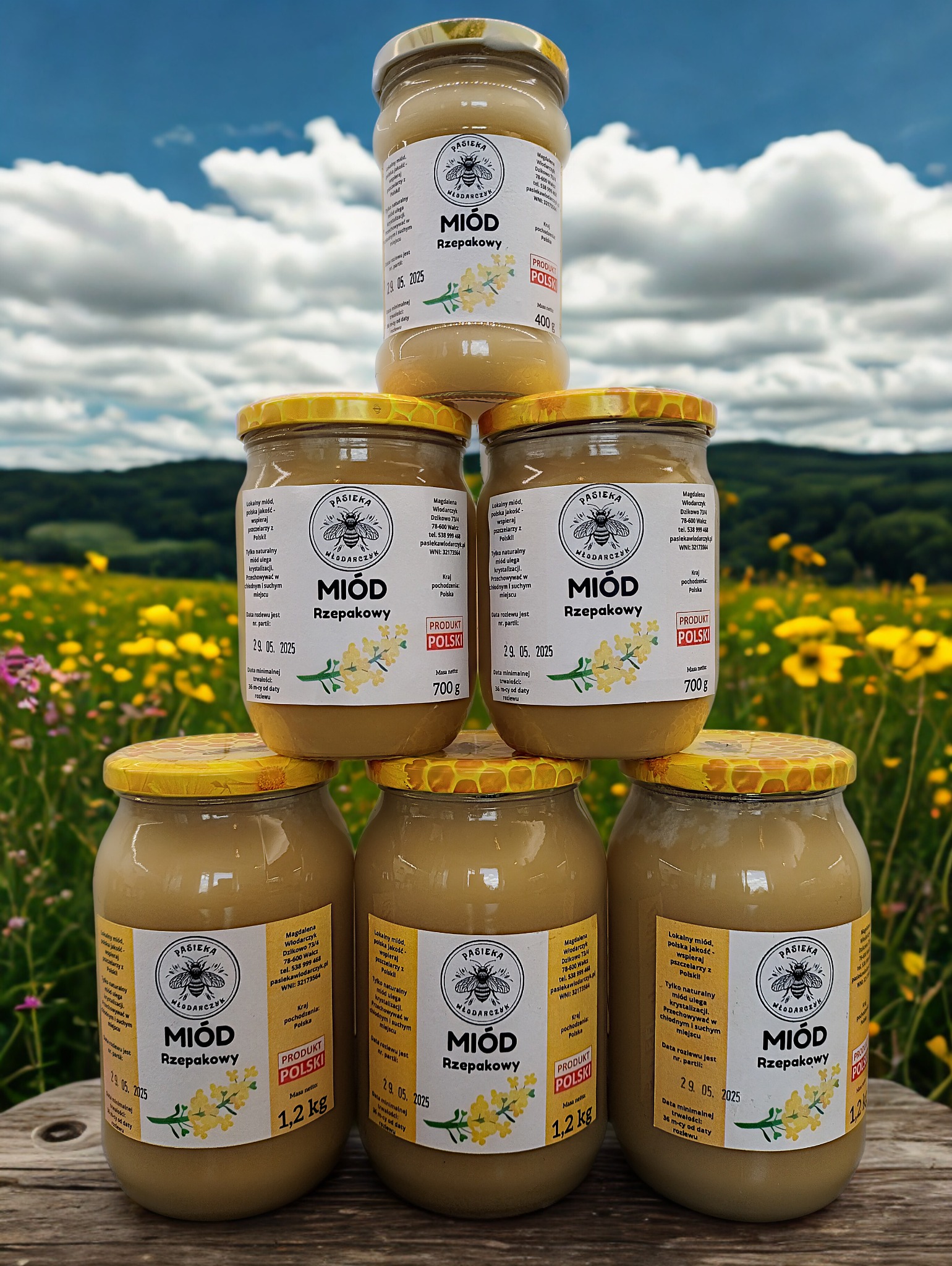 Piramida z słoików miodu rzepakowego Pasieki Włodarczyk na tle łąki i błękitnego nieba z chmurami. Widoczne słoiki o różnej gramaturze (400g, 700g, 1.2kg).
