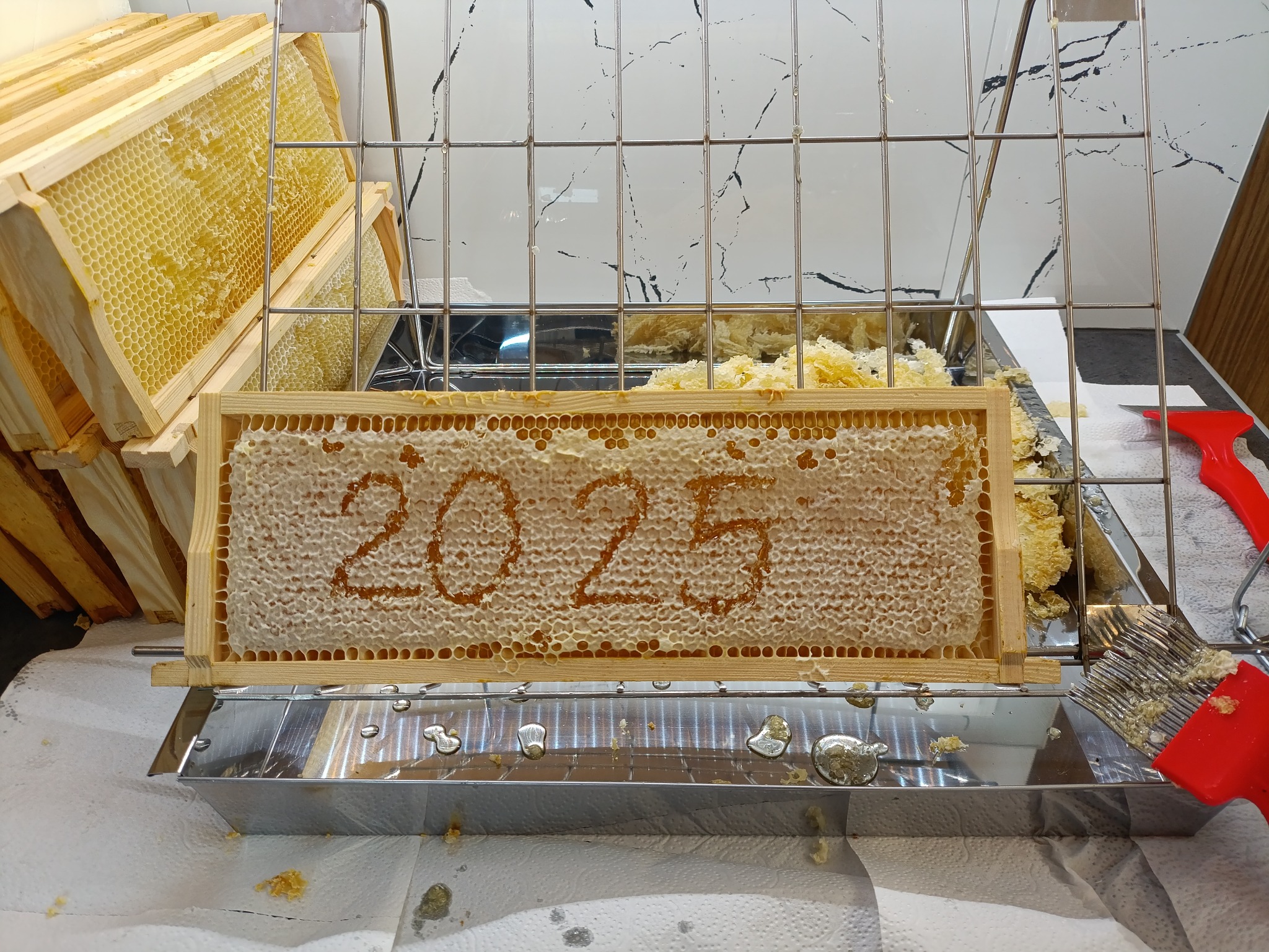 Ramka z plastrem miodu z wyciętym napisem '2025' w metalowej wytwornicy, obok narzędzia pszczelarskie i stos ramek. Koncept rocznicowy z produktem pszczelim.