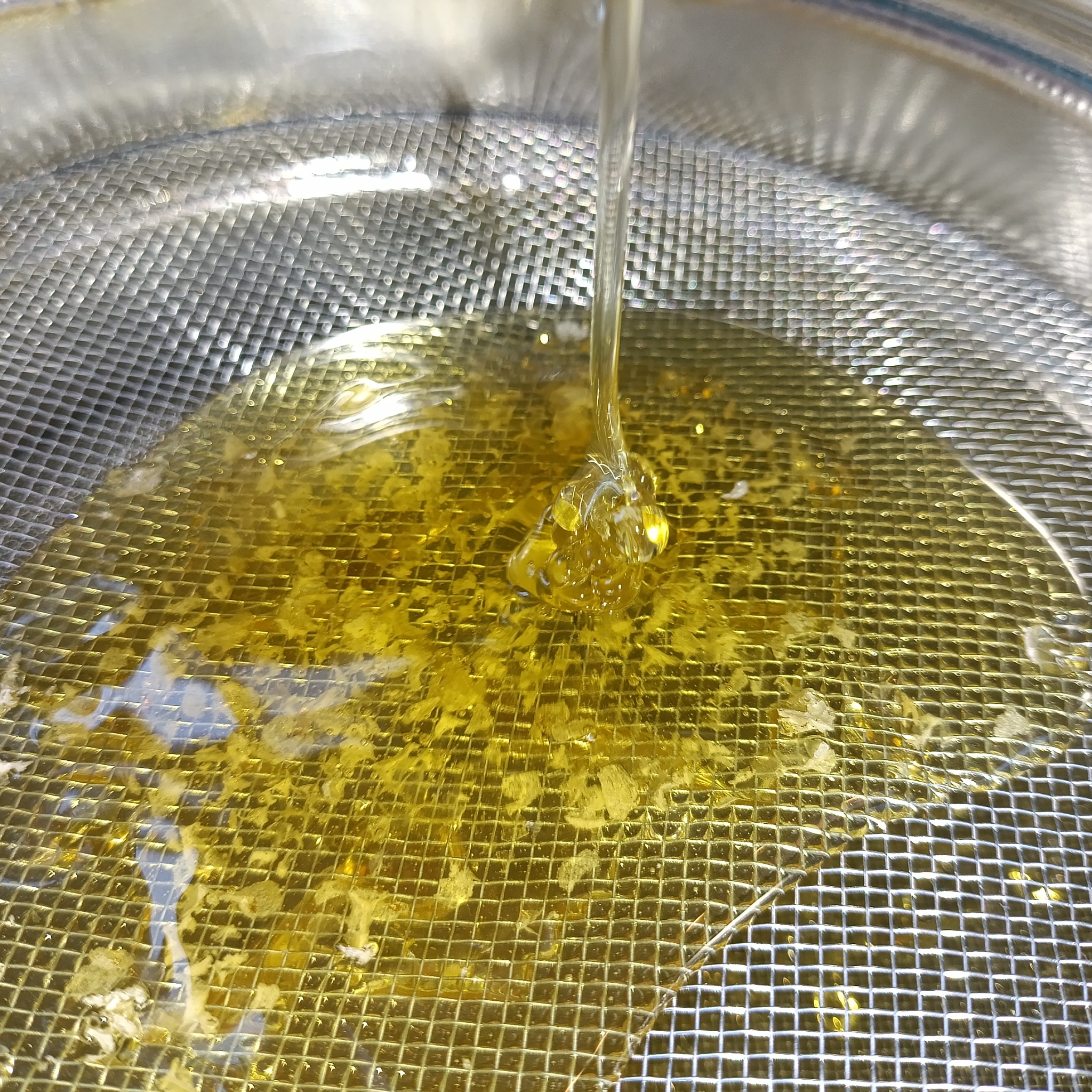 Proces filtracji świeżego, złotego nektaru przez metalowe sito. Ciecz spływa, tworząc delikatne krople i wzory na powierzchni.