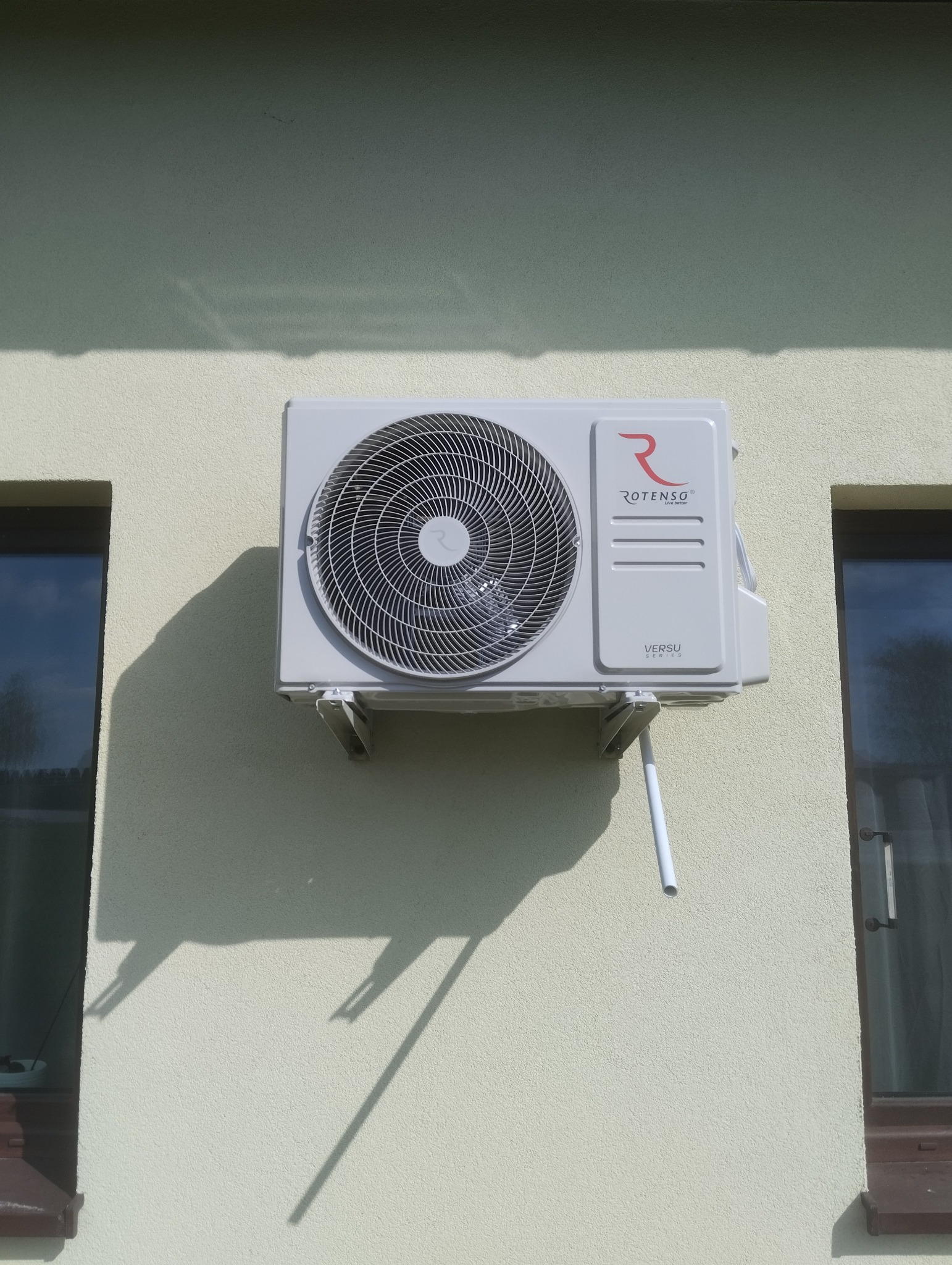 Zamontowana jednostka zewnętrzna klimatyzatora Rotenso Versu na elewacji budynku, widoczne okna po bokach, cień urządzenia na ścianie.
