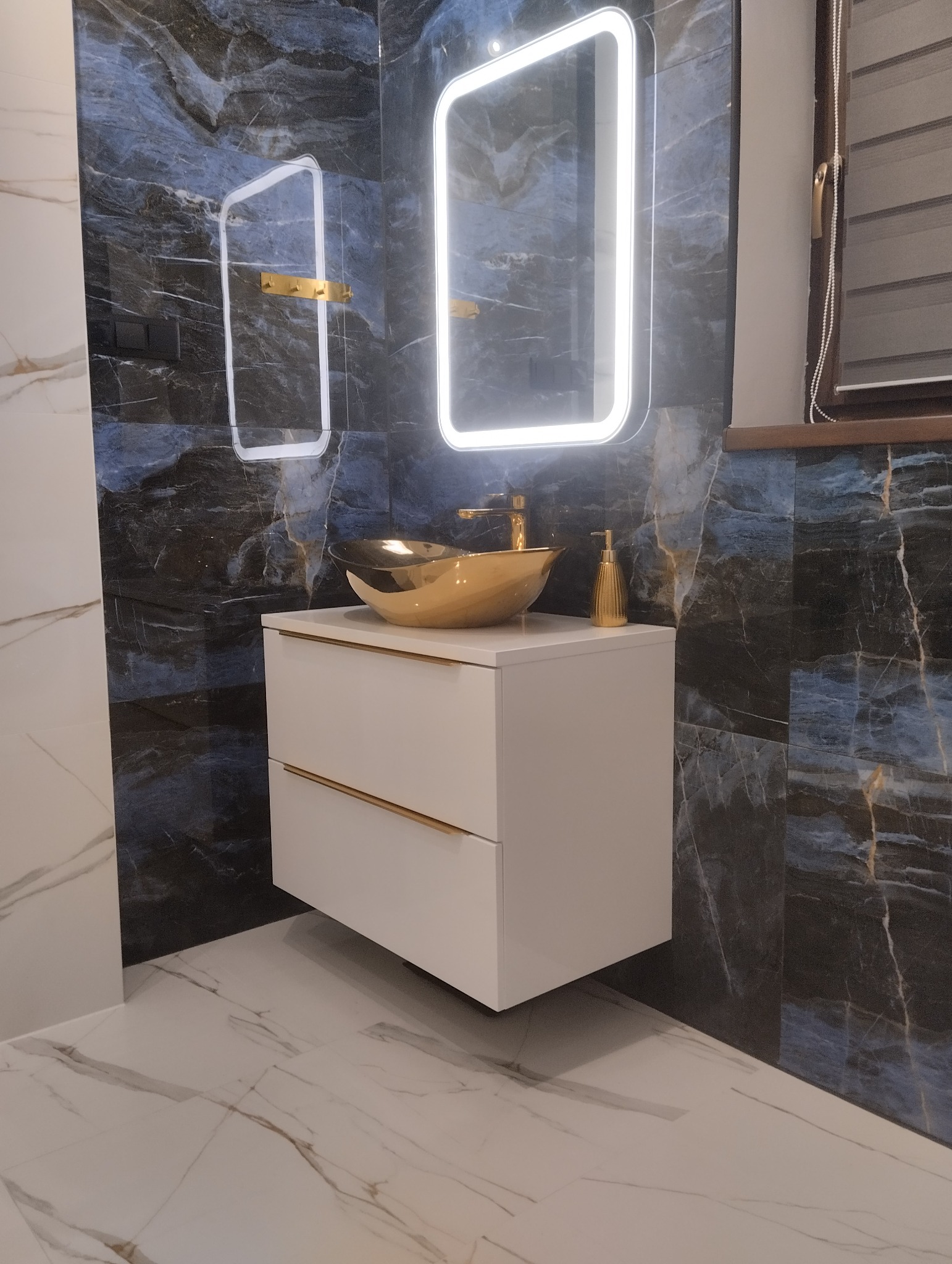 Nowoczesna łazienka z umywalką w kolorze złota, szafką z białymi frontami i podświetlanymi lustrami na ścianie z marmurowym wzorem.