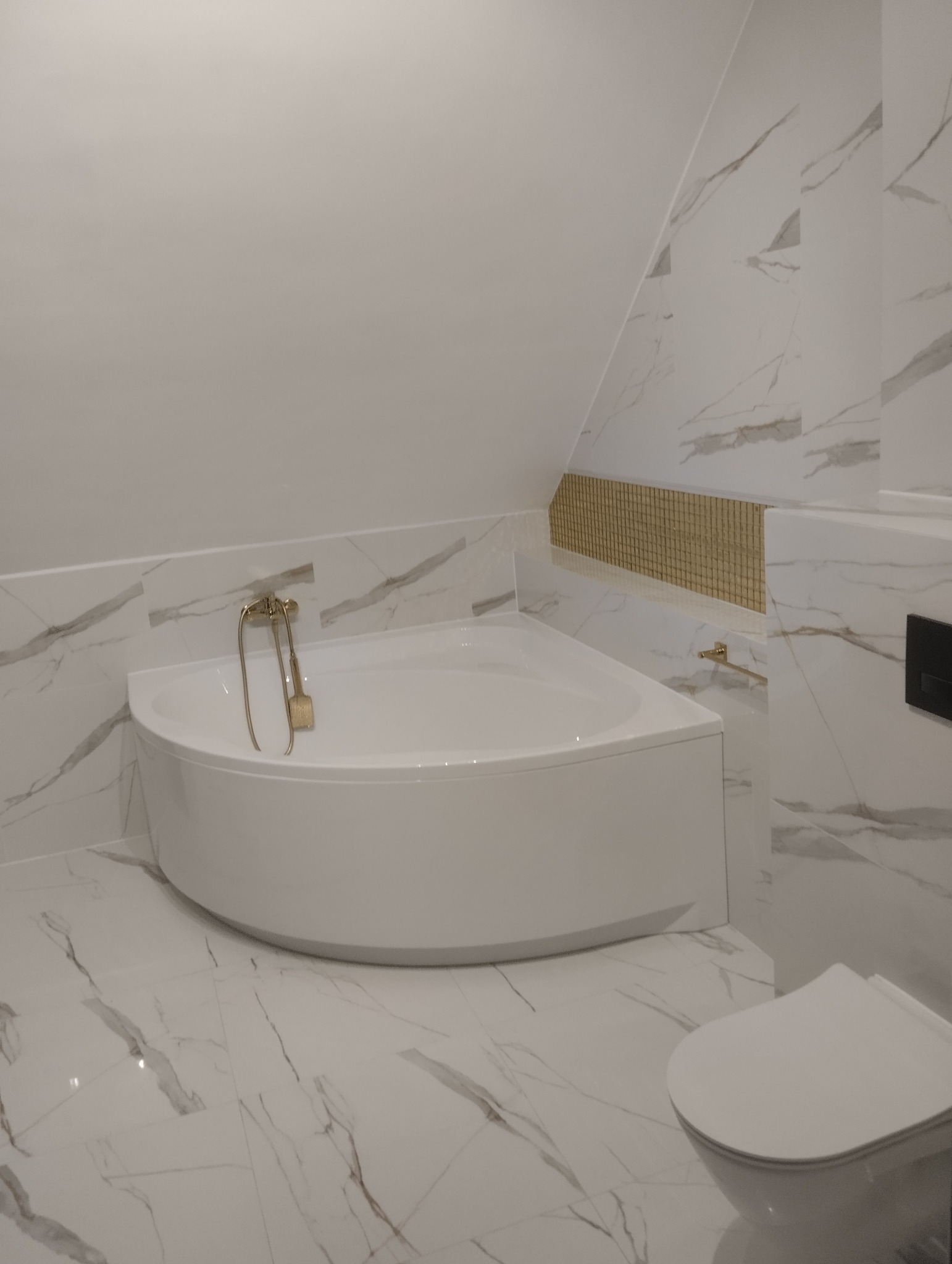 Nowoczesna łazienka na poddaszu z narożną wanną, złotą armaturą i marmurowymi płytkami.