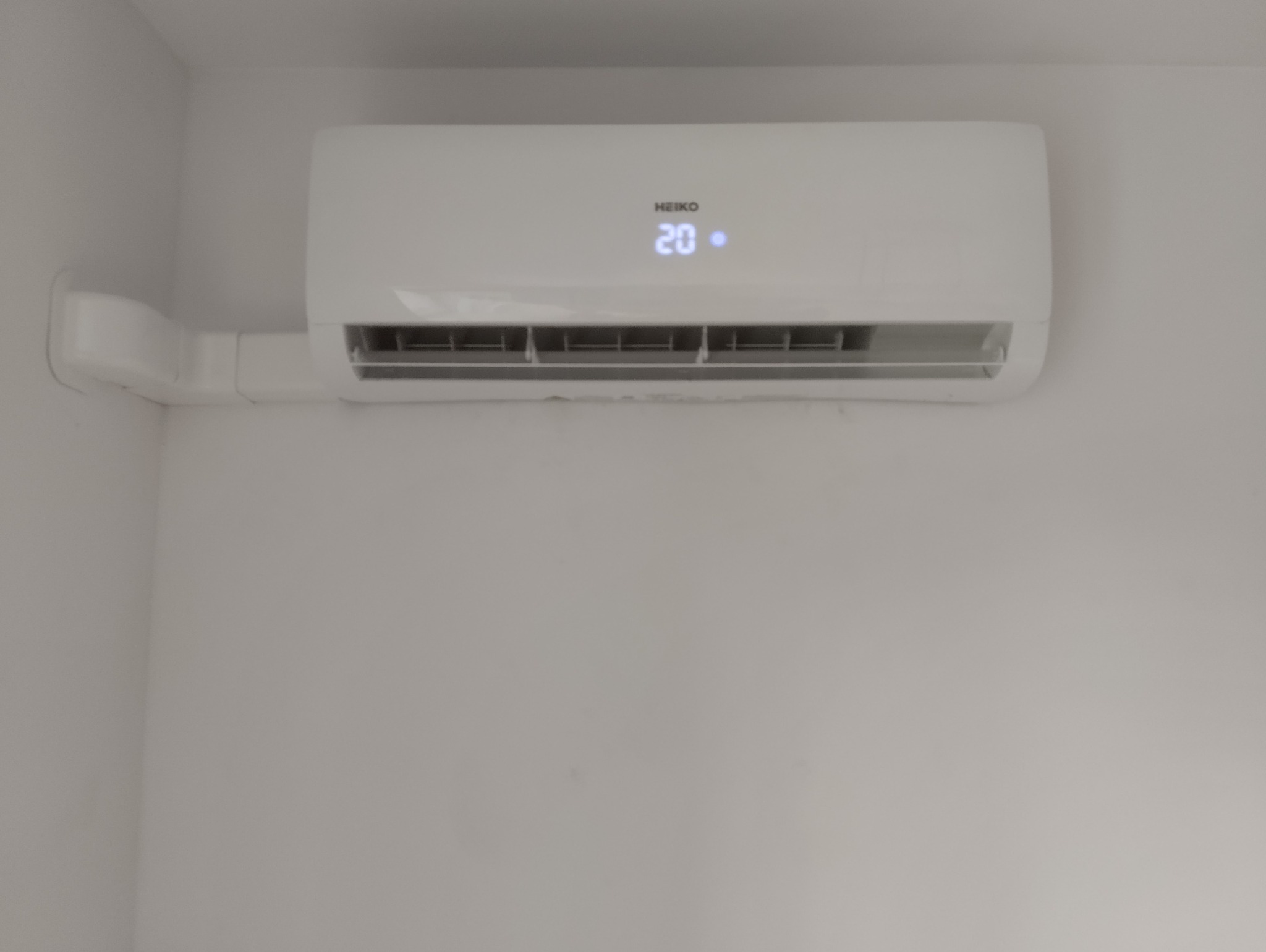 Biały klimatyzator HEIKO z wyświetlaczem temperatury 20 stopni Celsjusza, zamontowany na białej ścianie, z widocznymi przewodami w białej listwie.