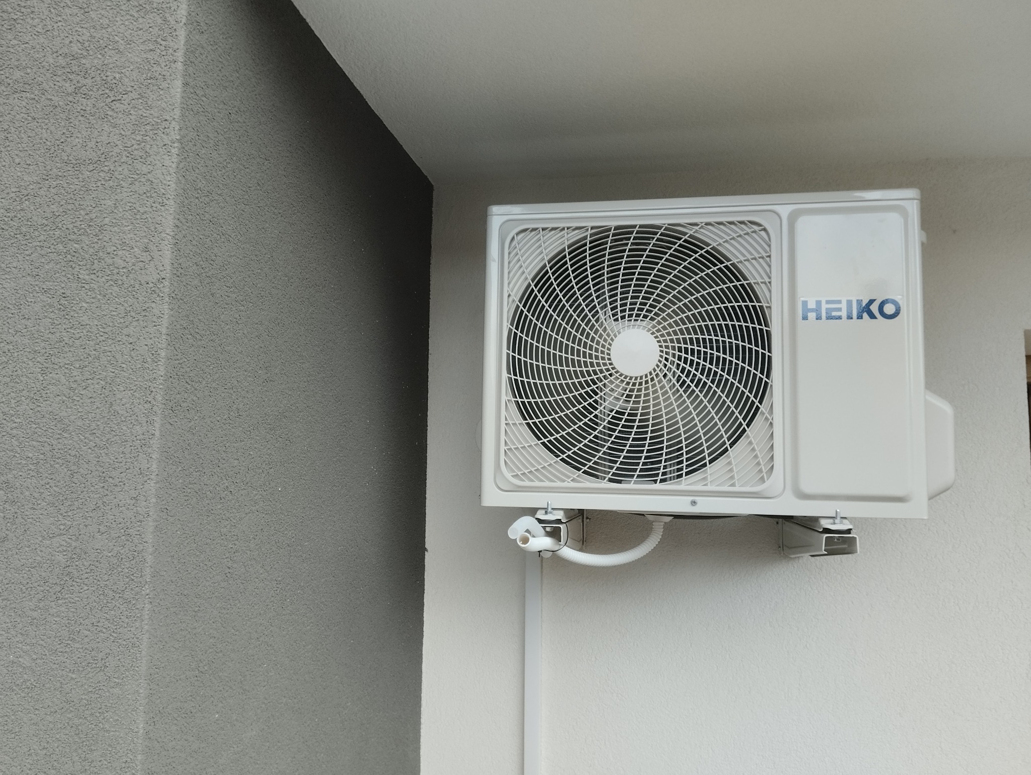 Biała jednostka zewnętrzna klimatyzacji marki HEIKO zamontowana na jasnej ścianie budynku z widocznymi elementami instalacji odprowadzającej skropliny.