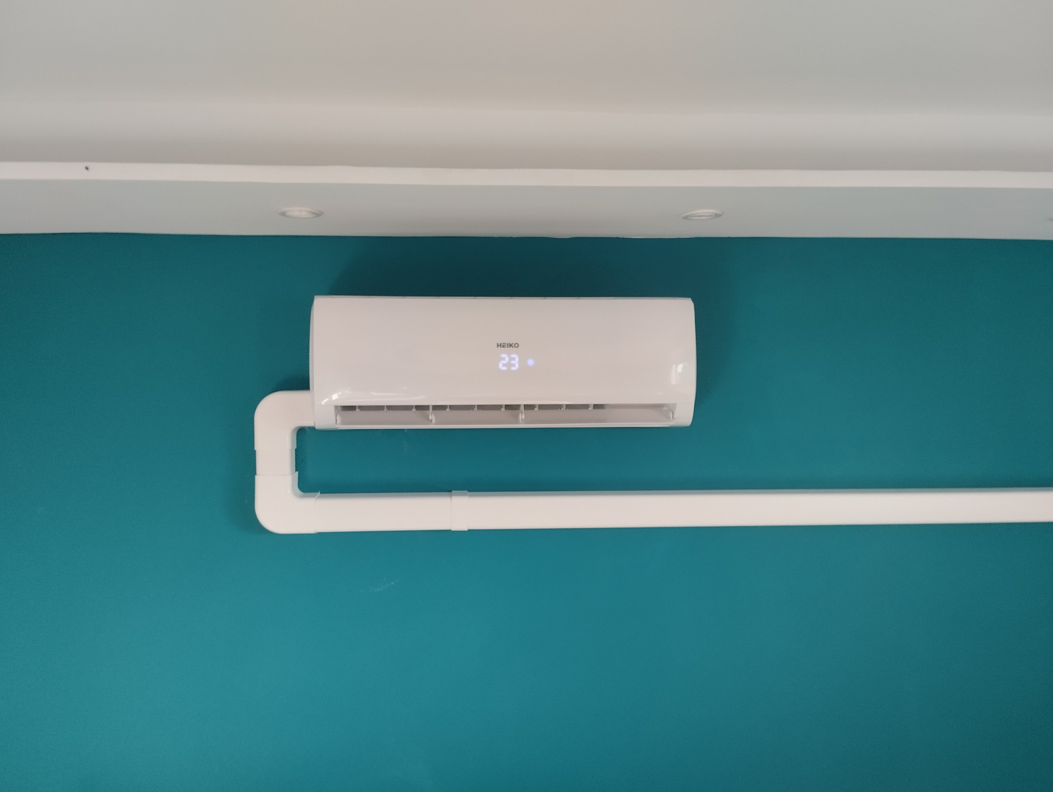 Biały klimatyzator HEIKO zamontowany na turkusowej ścianie, z widoczną instalacją odprowadzającą skropliny. Na wyświetlaczu widoczna temperatura 23 stopnie.