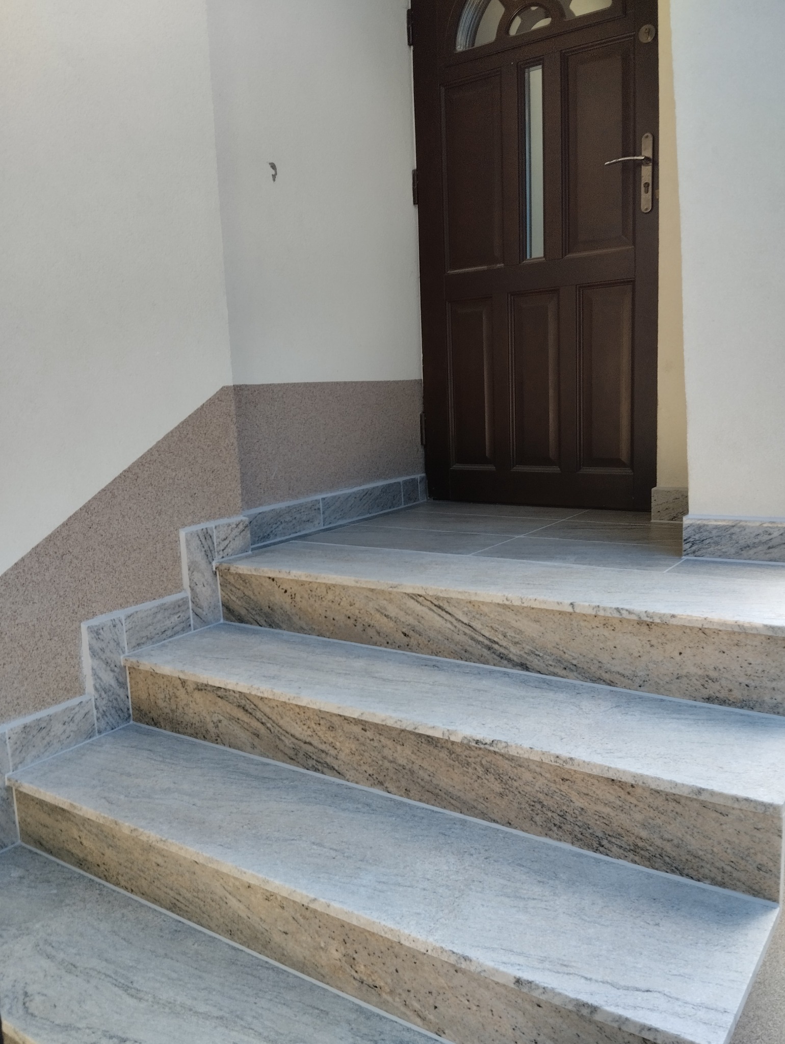 Wykończone marmurowe schody wejściowe prowadzące do brązowych drzwi z półokrągłym oknem. Ściana z dekoracyjnym tynkiem w dwóch odcieniach.