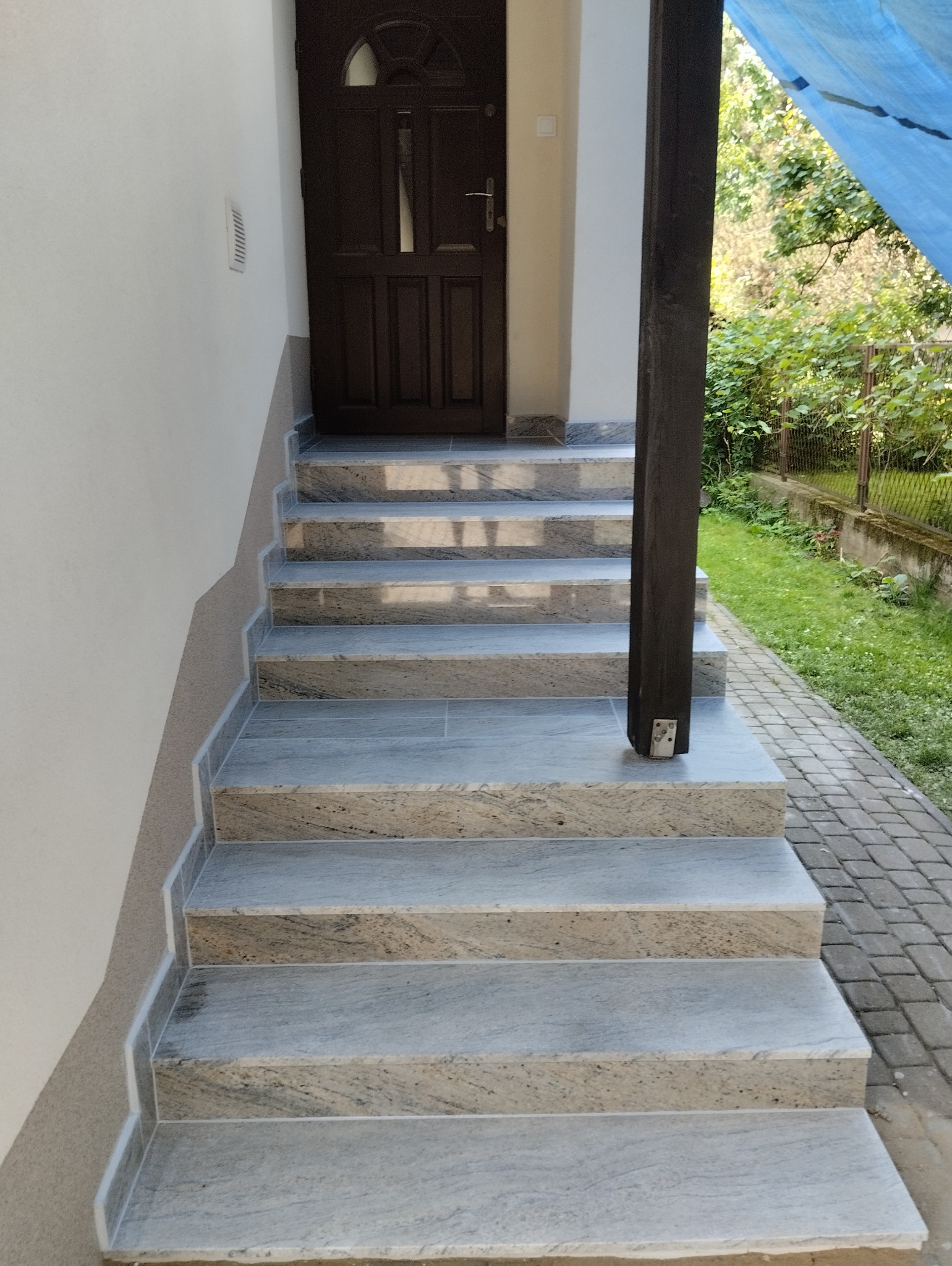 Granitowe schody wejściowe do domu, prowadzące do ciemnych drzwi. Widoczny fragment drewnianego słupa i kostka brukowa. Jasne barwy kamienia.