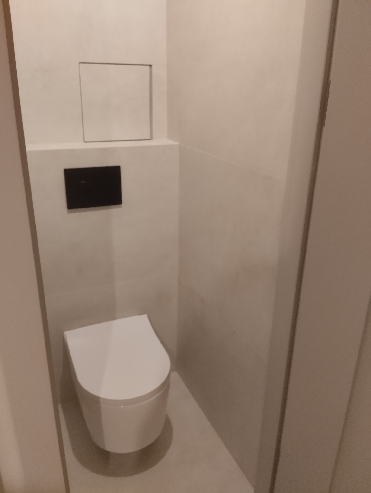 Nowoczesna toaleta podwieszana w minimalistycznej łazience z beżowymi płytkami, czarnym przyciskiem spłukującym i ukrytym dostępem rewizyjnym.
