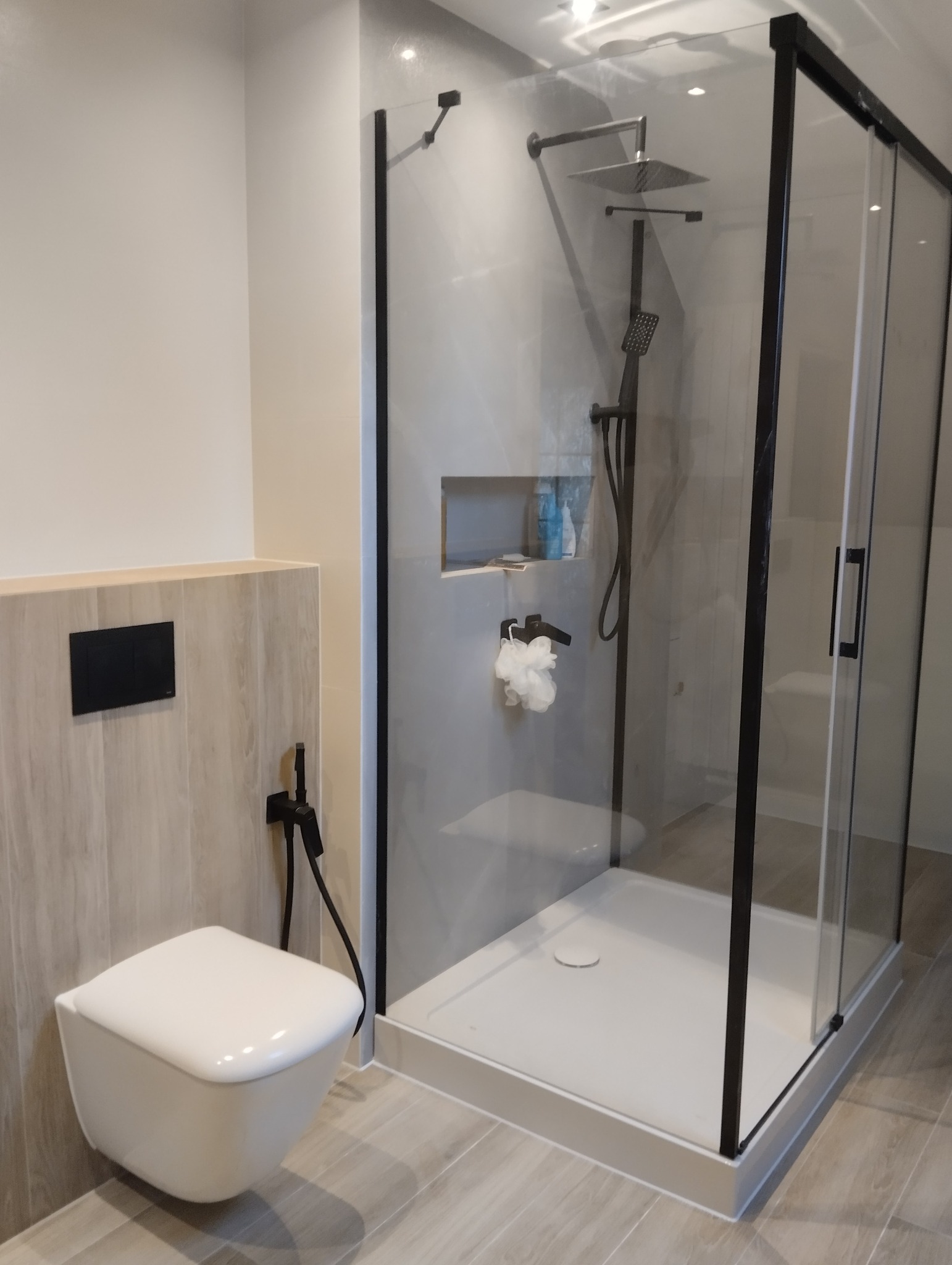 Nowoczesna kabina prysznicowa z czarnymi ramami i podwieszana toaleta w jasnej łazience z płytkami imitującymi drewno. Minimalistyczny design, oświetlenie punktowe.