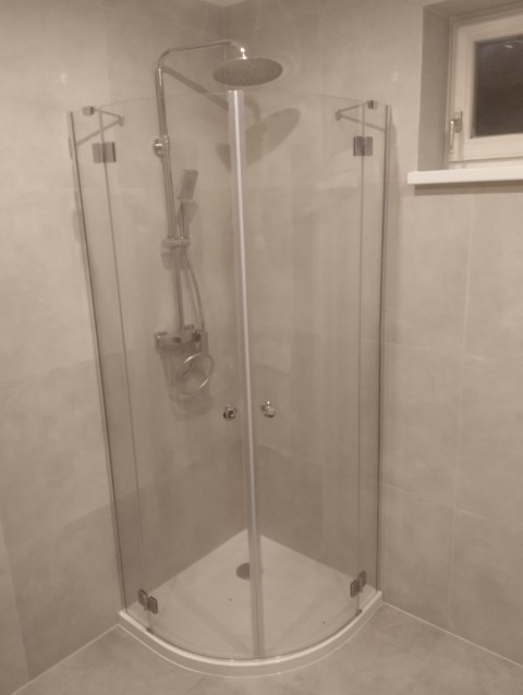 Narożna kabina prysznicowa z deszczownicą i szklanymi drzwiami, zamontowana w łazience wykończonej jasnymi płytkami. Widoczne okno z białymi ramami.