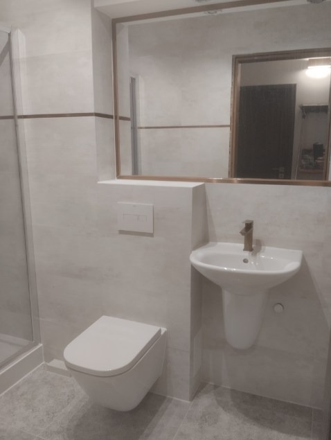 Nowoczesna łazienka z podwieszaną toaletą, umywalką na postumencie i dużym lustrem z miedzianą ramą. Minimalistyczny design, jasne płytki i szklana kabina prysznicowa.