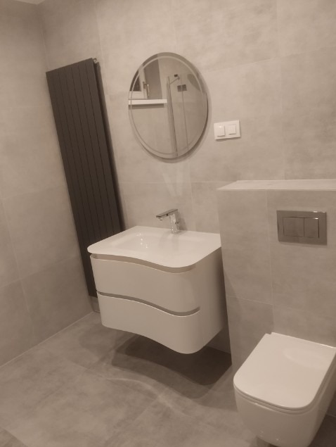 Nowoczesna łazienka w szarościach: podwieszana umywalka z szafką o falistym kształcie, owalne lustro i grzejnik drabinkowy. Minimalistyczny design, płytki imitujące kamień.