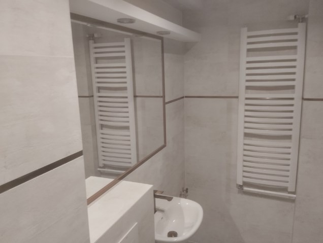 Biała łazienka z grzejnikiem drabinkowym, umywalką i lustrem. Widoczne płytki w odcieniach bieli i beżu z delikatnym wzorem. Minimalistyczny design.