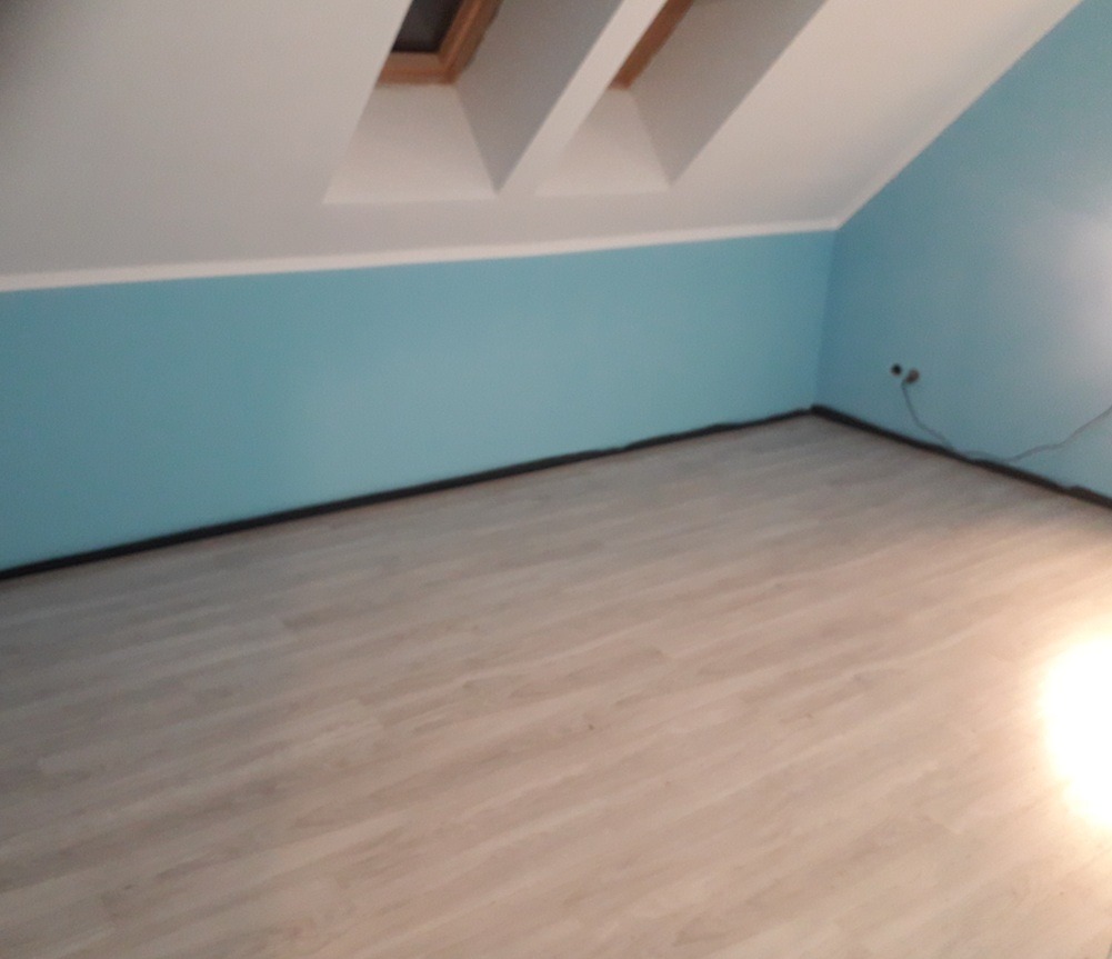 Pokój na poddaszu z panelami podłogowymi i błękitnymi ścianami. Widoczne okna dachowe i listwy przypodłogowe. Gniazdka elektryczne na ścianie.
