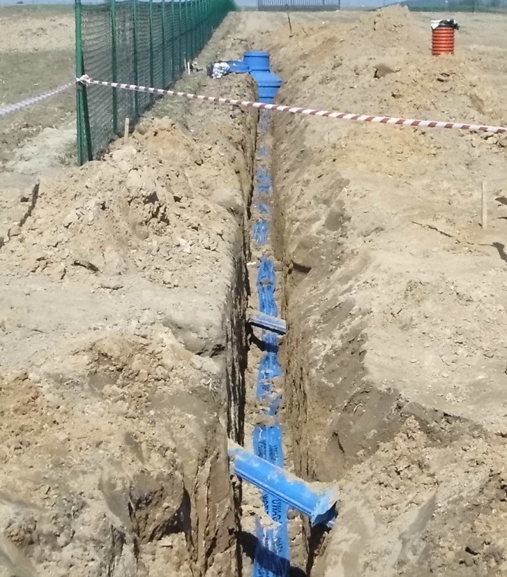 Wykop z ułożonymi niebieskimi rurami instalacyjnymi, zabezpieczony taśmą ostrzegawczą. Widok z góry na wykop w ziemi, z zielonym ogrodzeniem w tle.