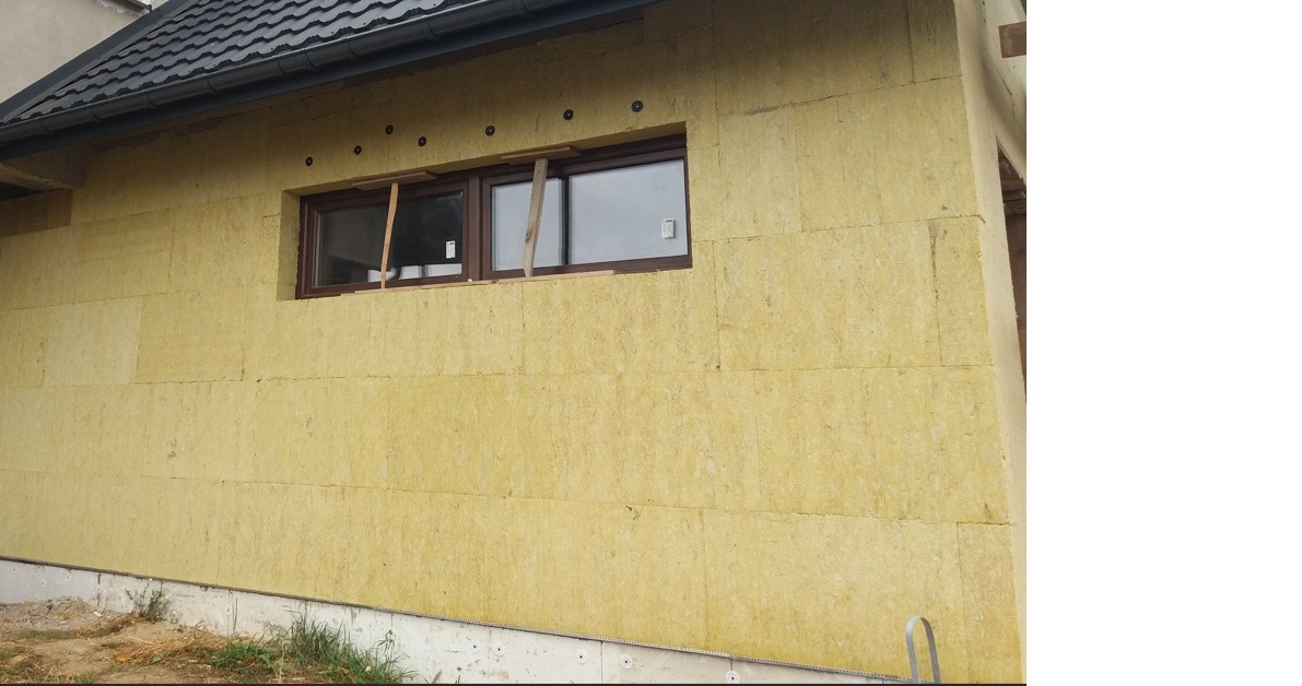 Ściana domu w trakcie ocieplania wełną mineralną, widoczne okno z drewnianą ramą i fragment dachu z czarną dachówką. Prace izolacyjne w toku.