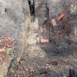 Darek - Głęboki wykop w ziemi, częściowo wyłożony cegłami, w tle widoczna osoba w roboczych ubraniach i kaloszach.