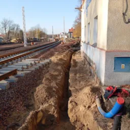 Wkop pod instalacje wzdłuż budynku dworca kolejowego, widoczne rury instalacyjne i fragment torów.