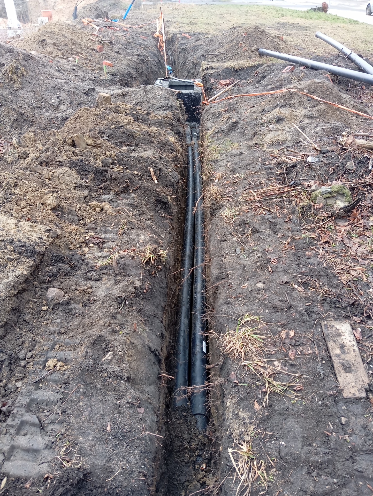 Wąski wykop w ziemi z ułożonymi dwoma czarnymi rurami, widoczny fragment studzienki rewizyjnej i pomarańczowa taśma ostrzegawcza.