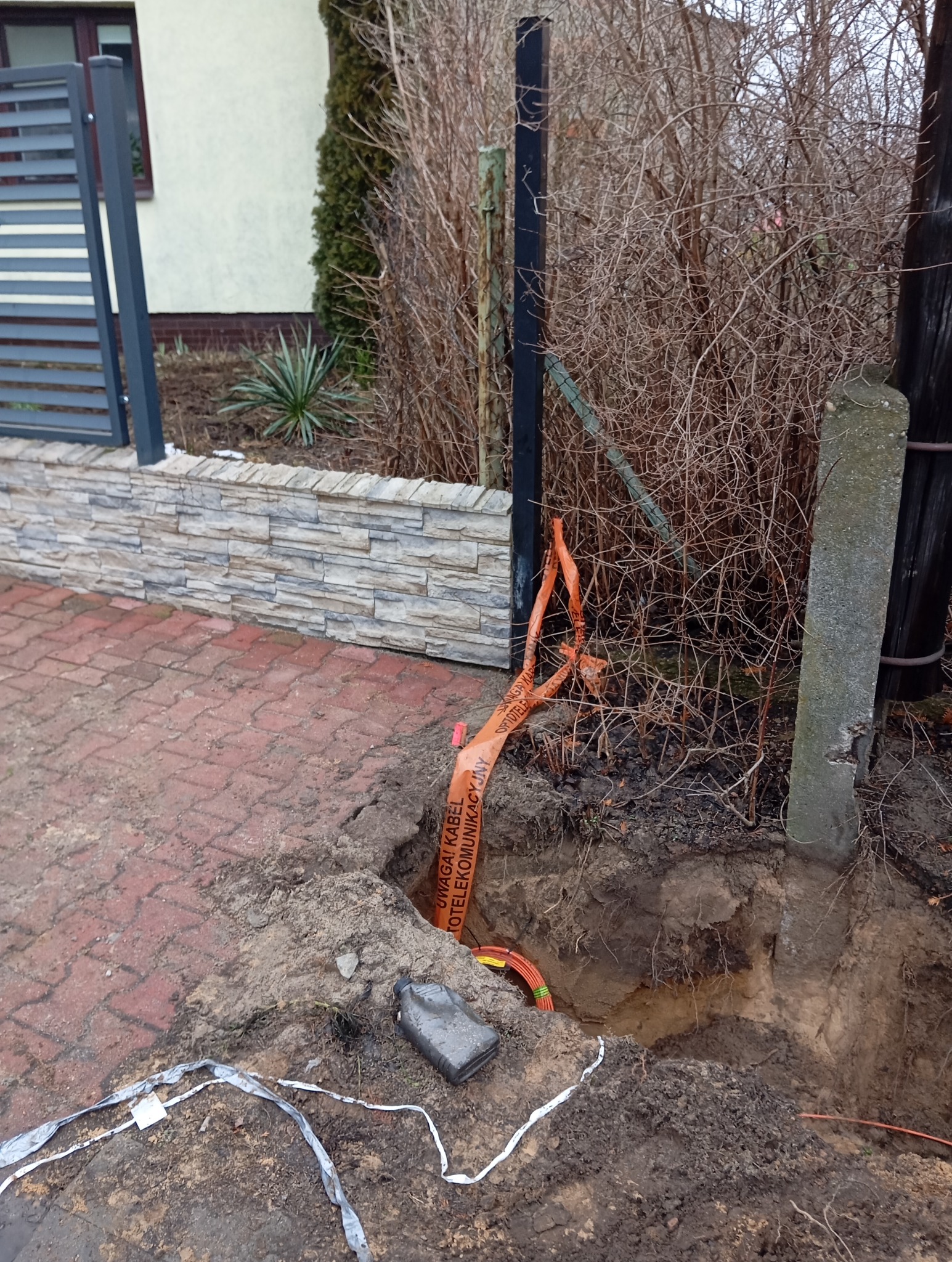 Wykop w ziemi z pomarańczową taśmą ostrzegawczą 'Uwaga! Kabel telekomunikacyjny', widoczne kable w wykopie, w tle ogrodzenie, krzewy i fragment budynku.