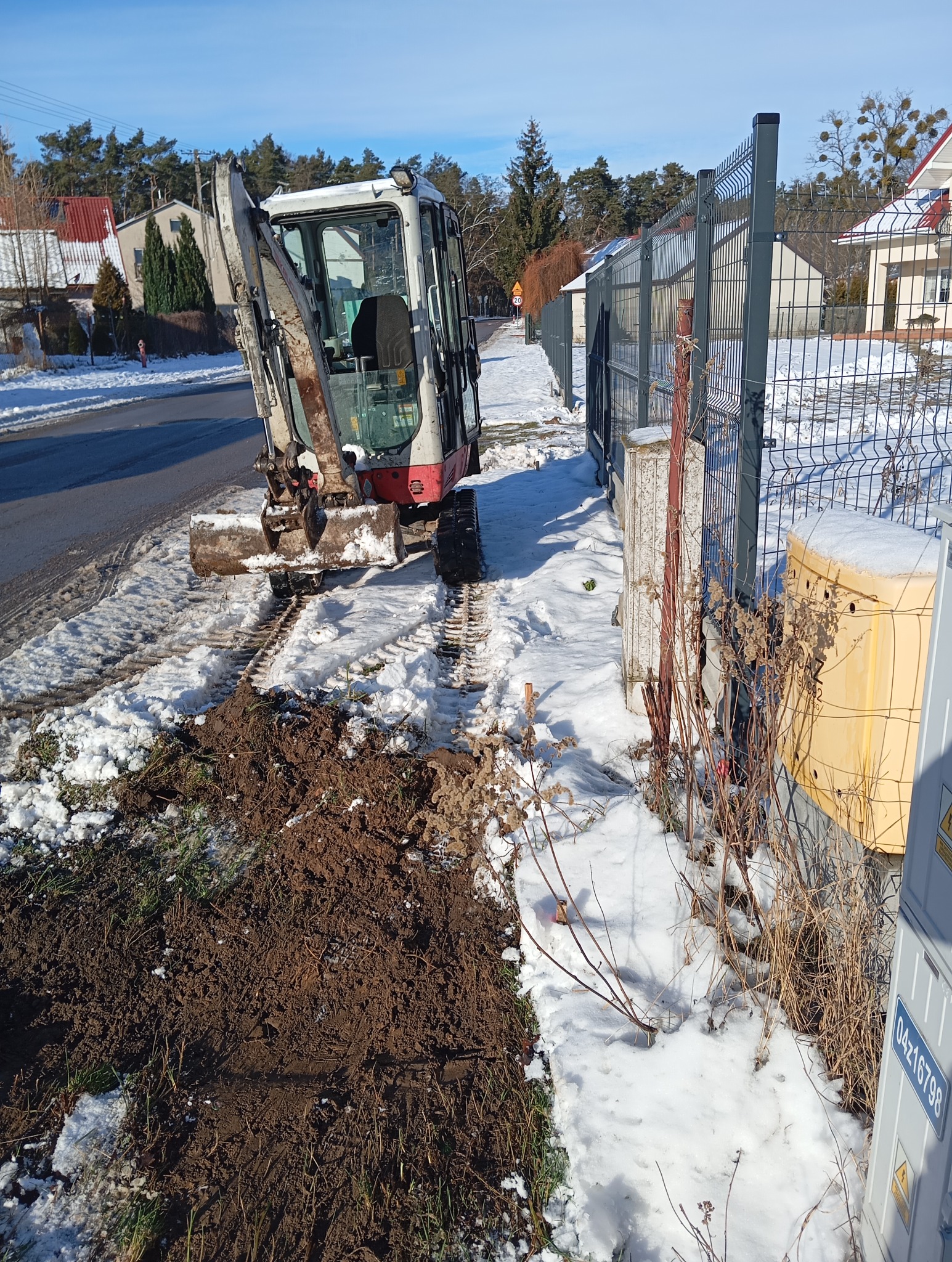 Mini koparka na śniegu obok wykopanego rowu wzdłuż ogrodzenia, z widocznymi śladami gąsienic i stertą ziemi.