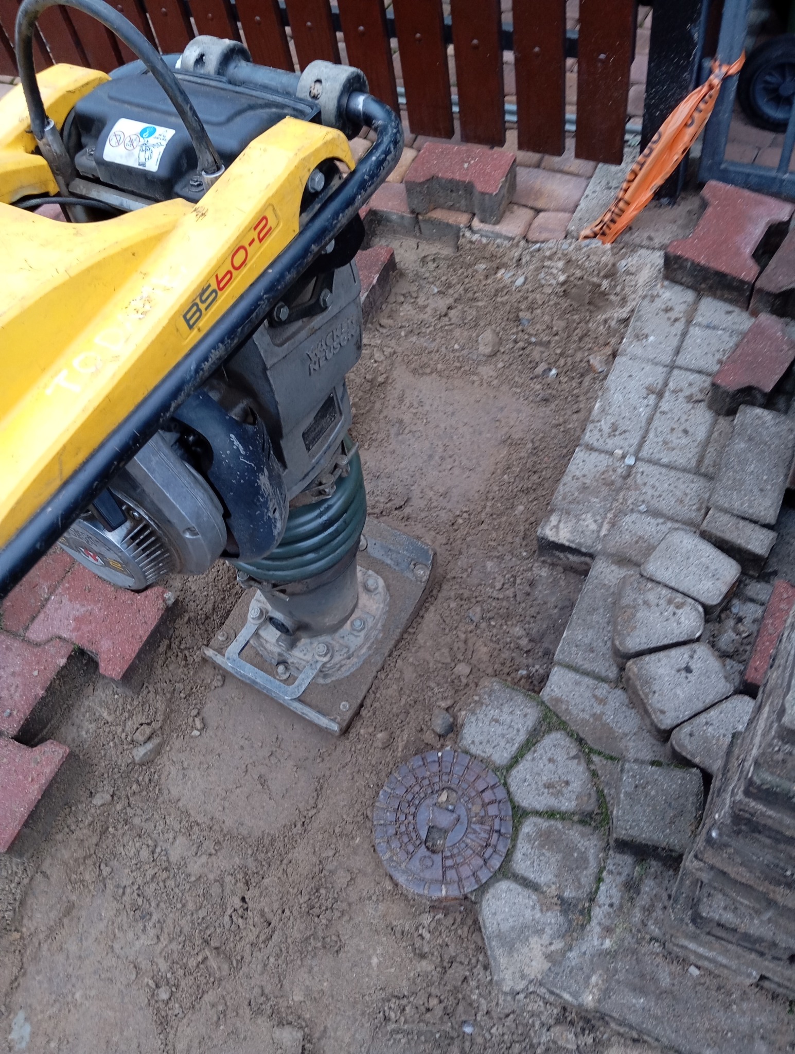 Żółta zagęszczarka Wacker Neuson BS60-2 ubija ziemię obok studzienki kanalizacyjnej i ułożonych częściowo kostek brukowych.