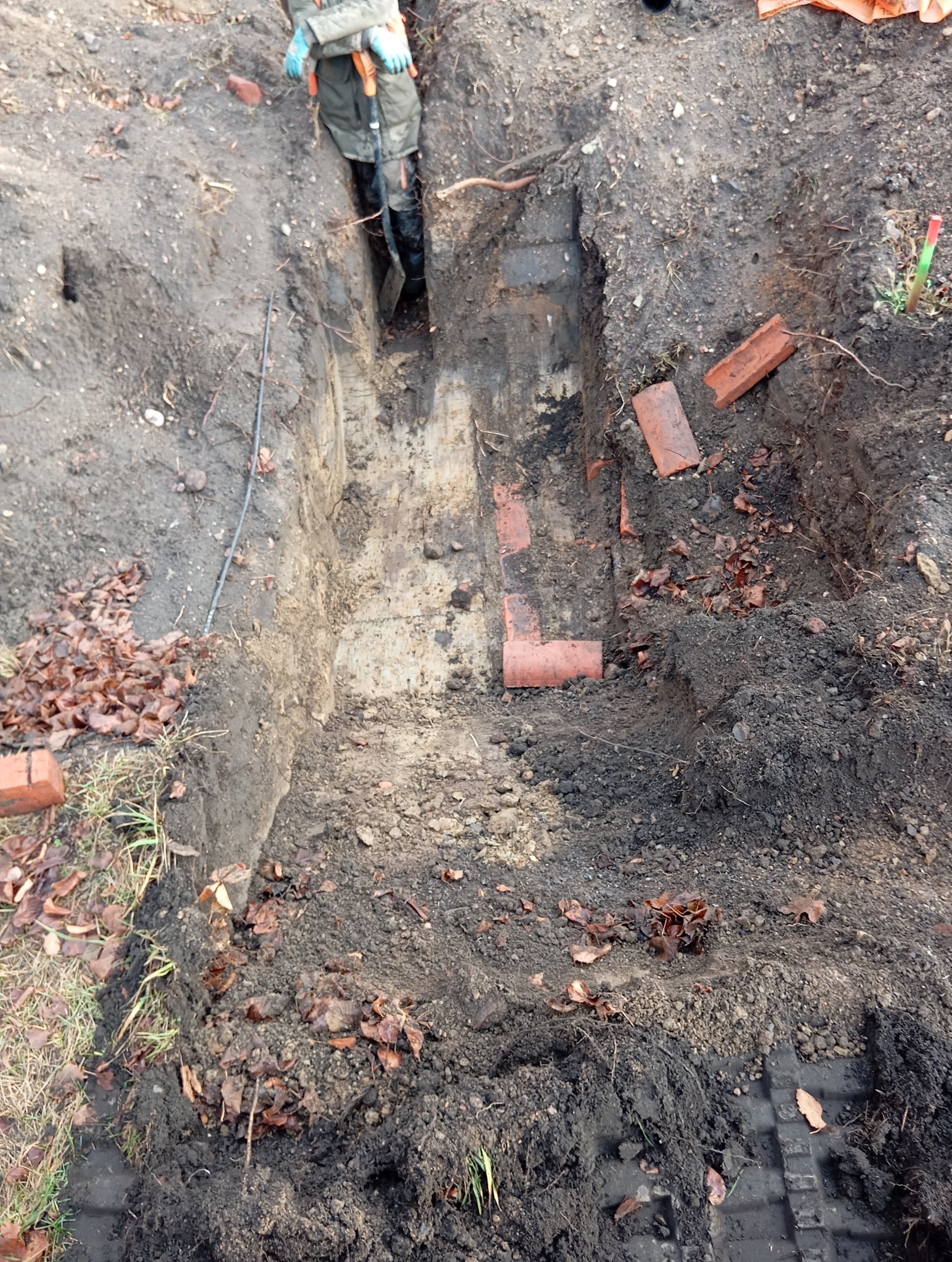 Głęboki wykop w ziemi, częściowo wyłożony cegłami, w tle widoczna osoba w roboczych ubraniach i kaloszach.